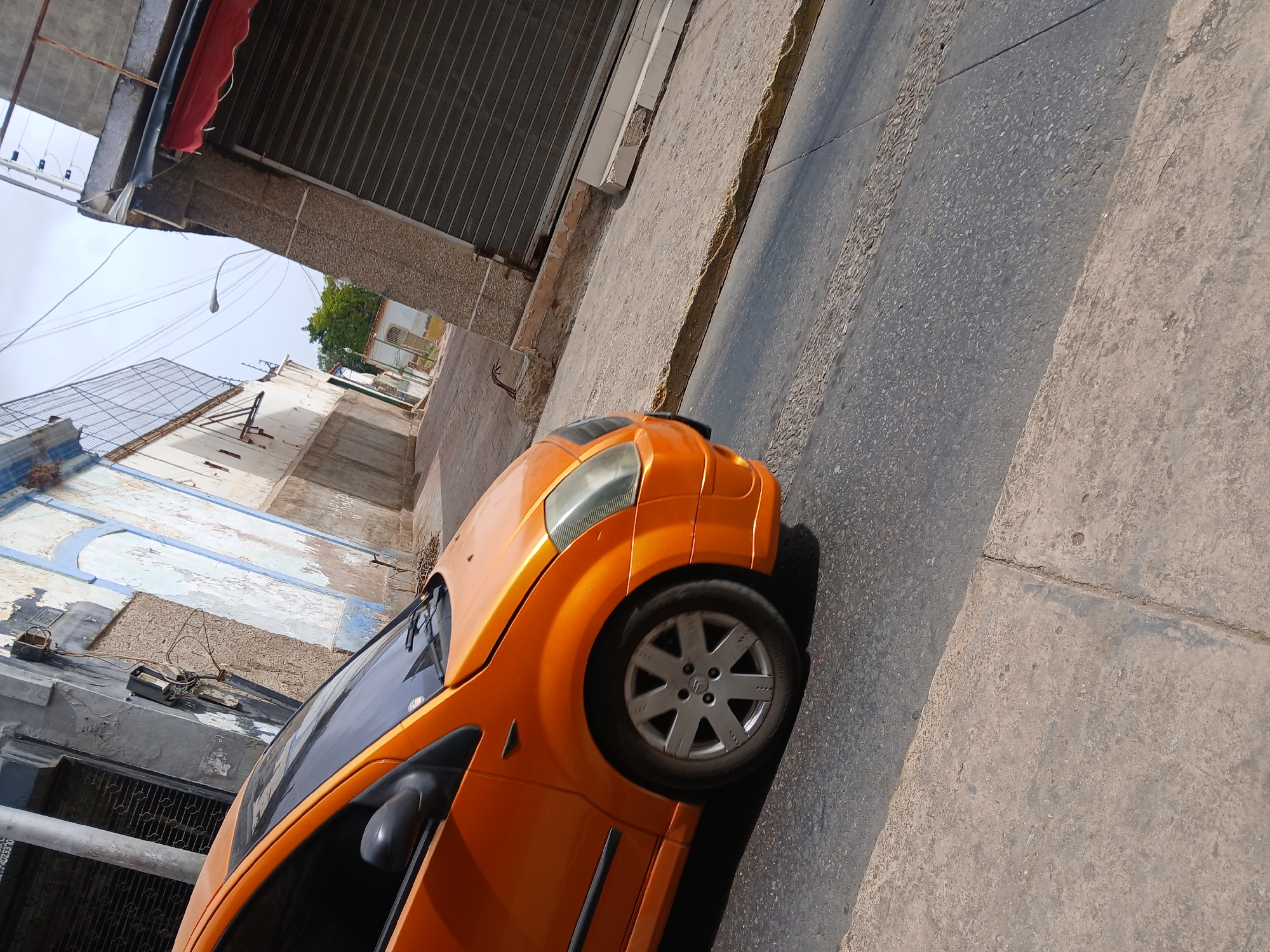 An orange Citroen C2 is parked on the side of a narrow street in Juan Griego, Venezuela. The street is paved with concrete slabs and asphalt. Buildings line the street, with some showing signs of age and disrepair. The architectural styles visible indicate it might be a historic part of the city. A few utility lines are also visible.