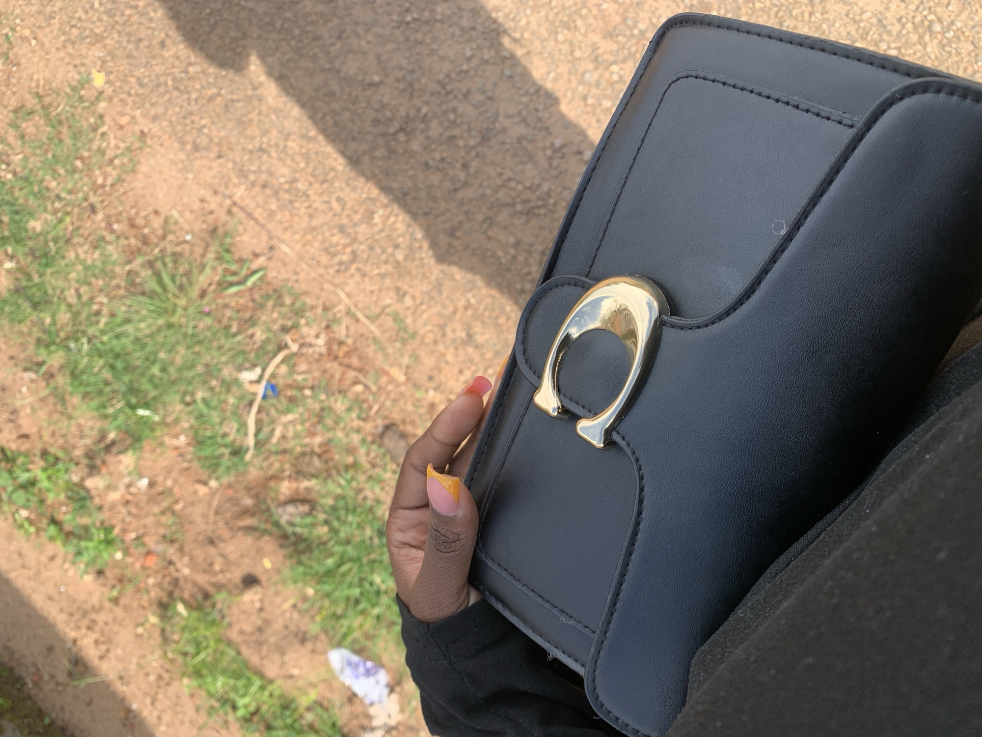 A first-person perspective shot shows a hand with bright orange and pink manicured nails resting on a black leather handbag. The handbag has a prominent gold buckle with a "C" shape. The background is out of focus, revealing a sandy, dirt path with patches of green grass and some litter, suggesting an outdoor urban or suburban environment. The lighting indicates it is daytime, with a shadow of a person falling across the ground from the right.
