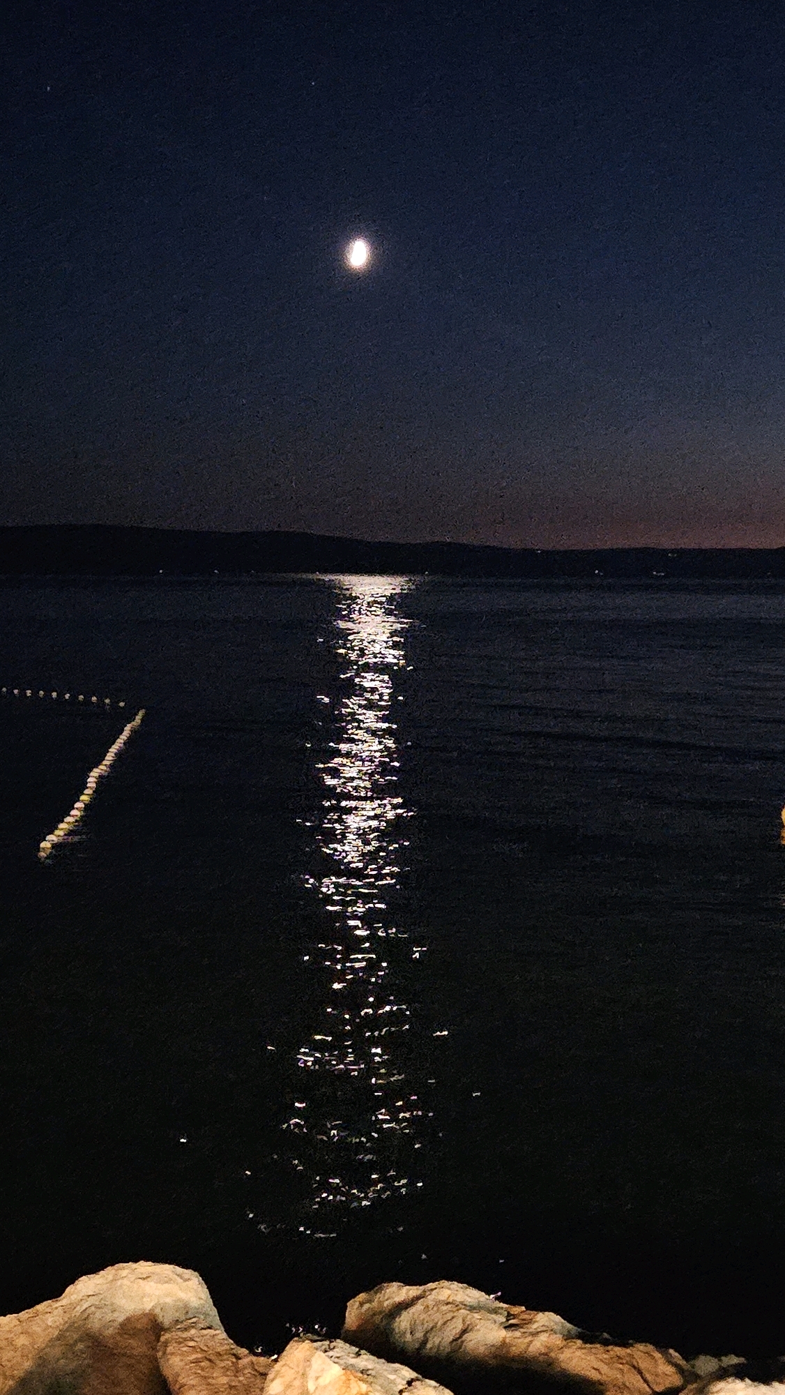A long exposure photograph captures a tranquil night scene by the Adriatic Sea in Općina Tučepi, Croatia. The moon, a luminous orb with a distinct crescent shape, hangs high in the deep blue, starry sky. Its reflection creates a shimmering, elongated path of light across the dark, rippling water. In the distance, the silhouette of a dark, hilly coastline stretches across the horizon, faintly illuminated by scattered lights that suggest the presence of a settlement. In the foreground, large, textured rocks are visible, their earthy tones contrasting with the deep blues and blacks of the night. Along the left edge of the water, a line of small, bright lights, possibly buoys or markers, stretches into the distance, adding a subtle linear element to the composition. The overall atmosphere is serene and contemplative, evoking the quiet beauty of a moonlit night on the Dalmatian coast.