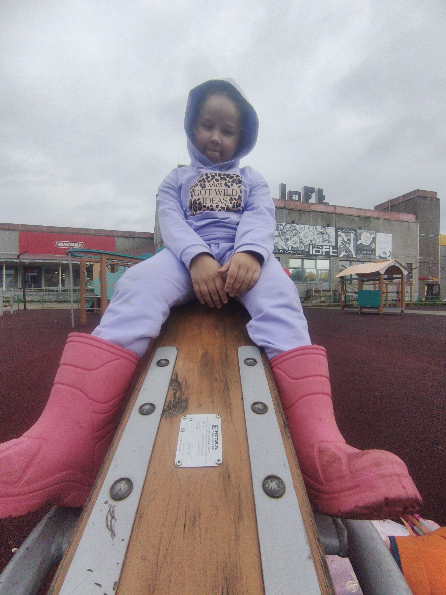 A young girl with dark curly hair sits on a seesaw, her legs extended in front of her and her feet clad in bright pink rubber boots. She is wearing a light purple hooded sweatshirt and matching pants. The sweatshirt has a graphic print on the chest that says "she's GOT WILD IDEAS" in leopard print lettering. The girl is looking down with a neutral expression.
The seesaw is made of wood with metal reinforcements. A sticker with text and a barcode is affixed to the wooden surface. The background shows a playground with a red rubberized surface and a few playground structures. In the distance, there are buildings, one of which has a red sign with the word "МАГНИТ" in white Cyrillic letters. Another building has the word "loft" in large letters above a storefront. The sky is overcast, suggesting a cloudy day. The overall scene appears to be an outdoor playground in a public area, possibly a shopping center or a recreational facility, in Markovsky, Russia. The atmosphere is subdued due to the cloudy weather.