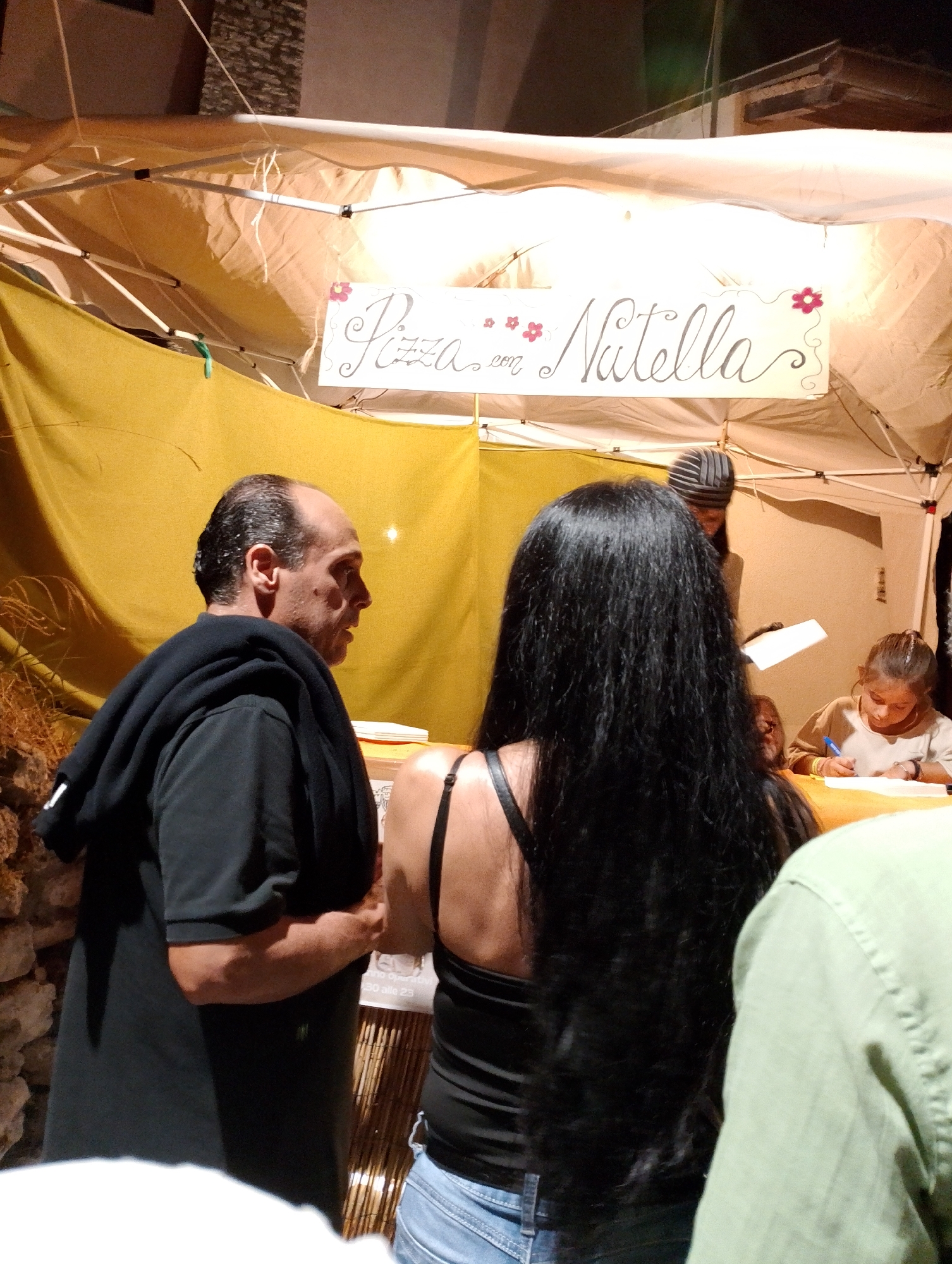 An outdoor market stall in Rivodutri, Italy, is depicted at night. A banner with "Pizza con Nutella" and floral decorations is displayed above. A man in a black hoodie stands on the left, facing right in profile. To his right, a woman with long black hair faces away from the camera. Behind them, another woman with braided hair is seated and writing, with a man in a striped hat behind her. The stall is supported by a white tent structure, with yellow fabric draped behind. A stone wall is visible on the far left.