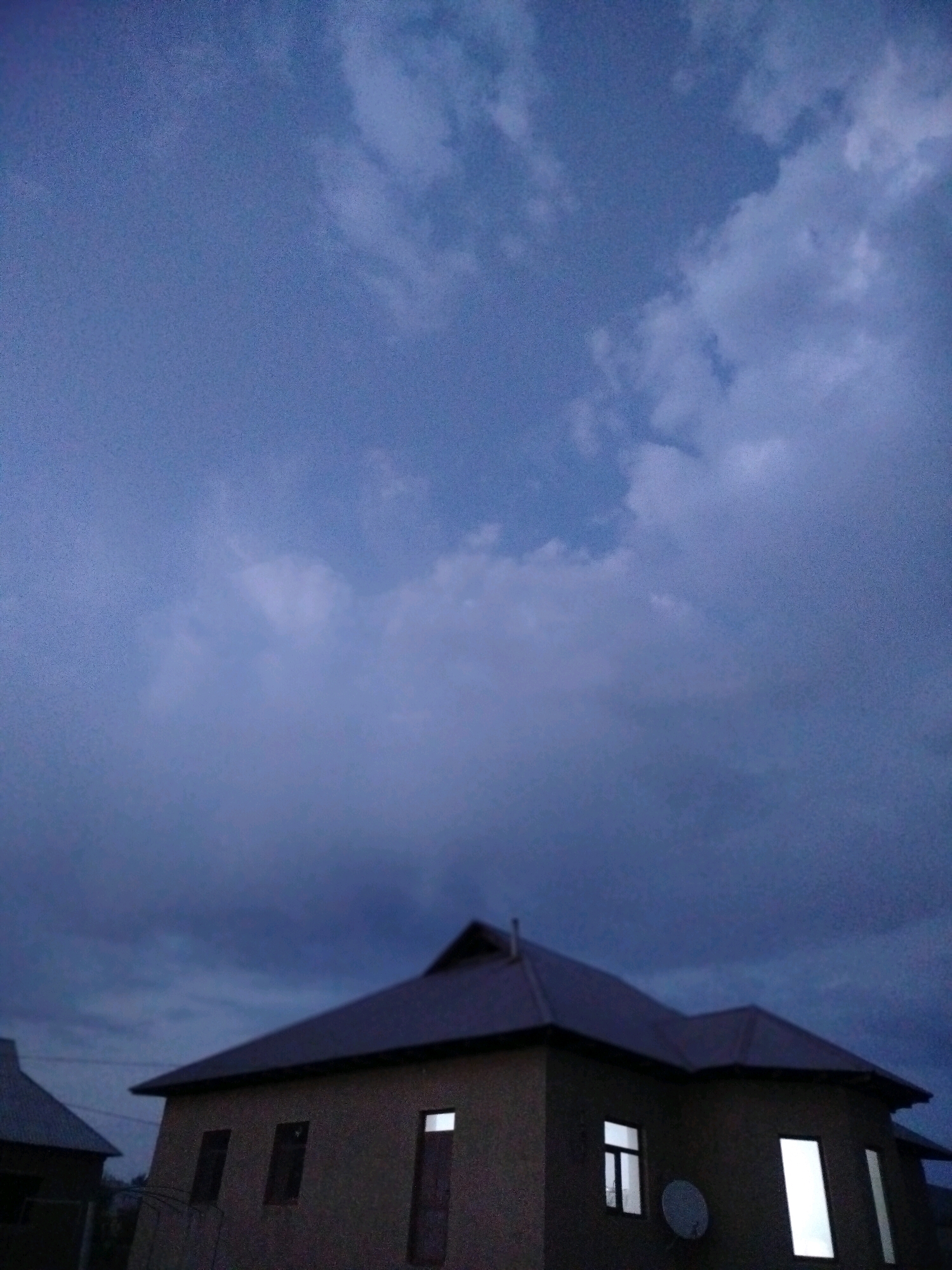 The image depicts a two-story house with a dark, sloped roof against a cloudy, dusky sky. The house has several windows, some of which are illuminated from within. A satellite dish is attached to the side of the building. To the left of the main house, a portion of another building with a similarly sloped roof is visible. The scene appears to be in a residential area, possibly on the outskirts of the city of Көлкент, Kazakhstan, given the architectural style and the natural elements. The lighting suggests it is either early morning or late evening.