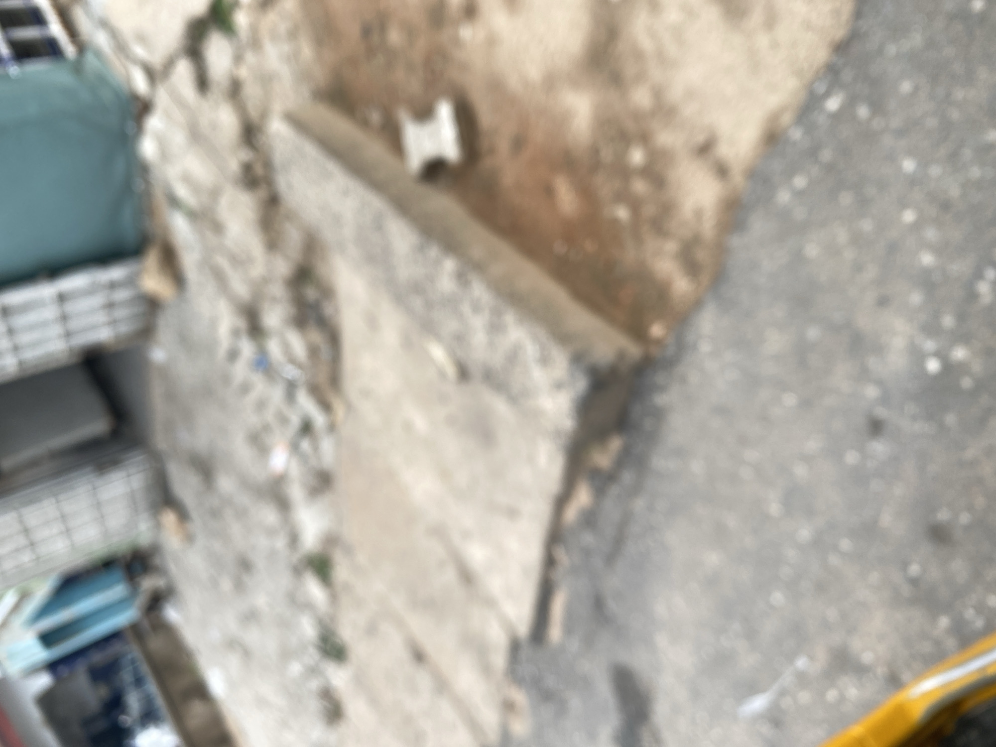 The image, taken in Jalingo, Nigeria, is a blurry, high-angle shot of an outdoor scene. The primary subjects appear to be concrete structures and debris on the ground. To the left, there are hints of what could be stacked crates or furniture, some covered in a teal fabric. Below this, metal shelving is visible.
The main focus is a textured, rough concrete surface that forms a ledge or a raised area. On this surface, a small, white, rectangular object, possibly a piece of plastic or debris, is noticeable. The ground around this concrete structure is a mix of dirt, gravel, and scattered refuse, with some greenery interspersed. The texture of the ground suggests it's an unpaved or neglected area.
The lighting is diffuse, making it difficult to determine the exact time of day, but it doesn't appear to be harsh sunlight. The overall impression is one of an urban or semi-urban environment, possibly a backyard, a construction site, or an alleyway. There is no discernible activity taking place; the scene is static. No text is visible in the image. The blurriness of the image prevents any clear identification of specific objects or details, and it’s impossible to infer emotions or specific weather conditions beyond what can be gathered from the general appearance of the environment.
