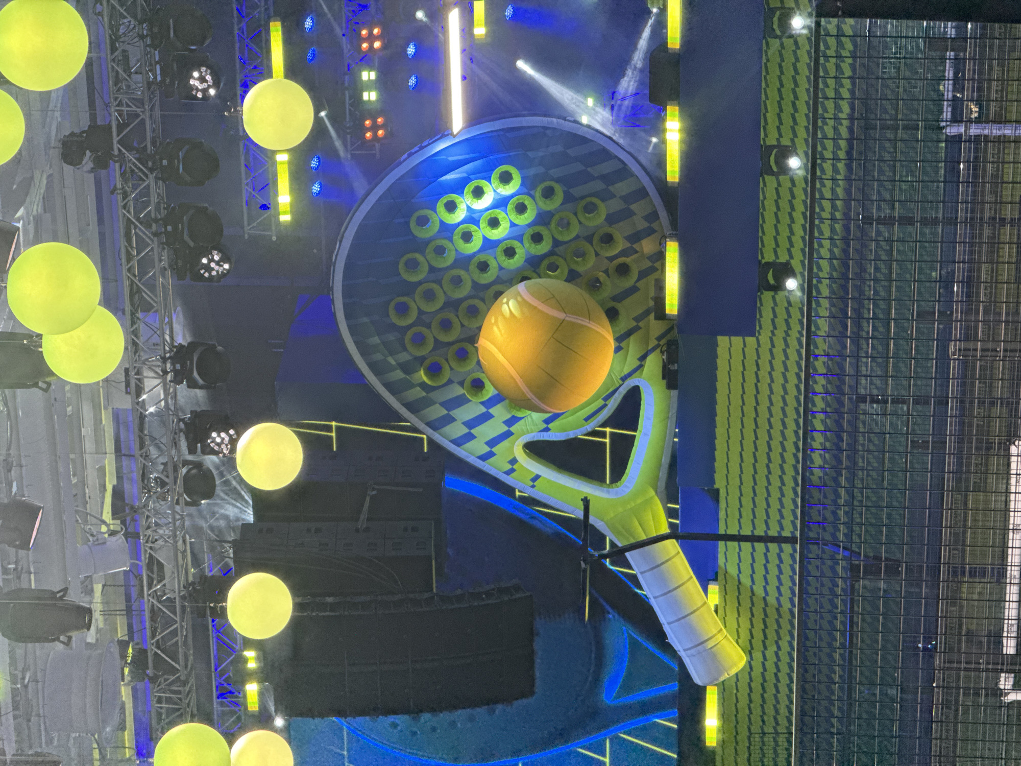 The image depicts an indoor scene, likely a stage or set design, featuring a large-scale decorative paddle tennis racket with a tennis ball in the center. The racket is primarily blue, yellow, and green, with a pattern of circles on the head and a white-striped handle. The tennis ball is yellow.
The background includes stage lighting fixtures, truss structures, and large, illuminated spheres hanging in the air. A wire fence is visible on the right side of the image.
Based on the information provided, this scene is located in Yekaterinburg, Russia.