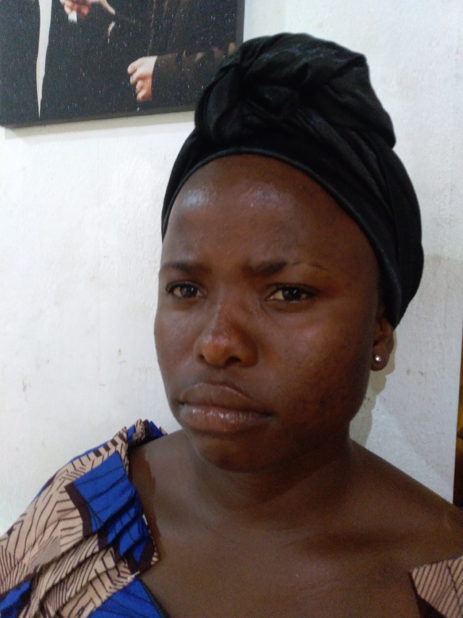 A medium close-up portrait captures an adult individual with dark skin, facing forward and looking directly at the camera. The subject is wearing a black headwrap and a garment featuring a blue, beige, and brown geometric pattern visible on the left shoulder. A silver stud earring is present in the visible right earlobe. The subject's facial expression is neutral, with lips slightly parted. The skin exhibits some textural variations and areas of reflectivity. The background consists of a light-colored, textured wall. In the upper left background, a framed picture or canvas is partially visible, depicting elements of two figures in dark formal attire with a hand visible centrally. The location is Unknown, Unknown.