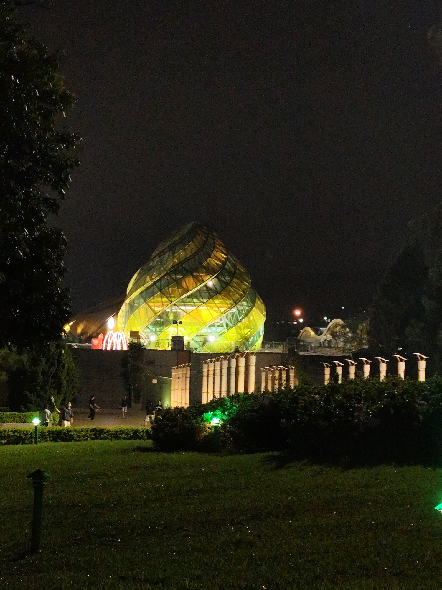 At night, a yellow-lit glass-covered structure, possibly a building, dominates the view. The structure is located behind a concrete barrier with white vertical columns. In the foreground, there is a green lawn with sparse lighting and shadowy bushes. The sky above is dark. People appear to be playing badminton on the grass area, lit by a lamp. This is likely the architecture of the Da Lat Flower Garden in Da Lat, Vietnam.