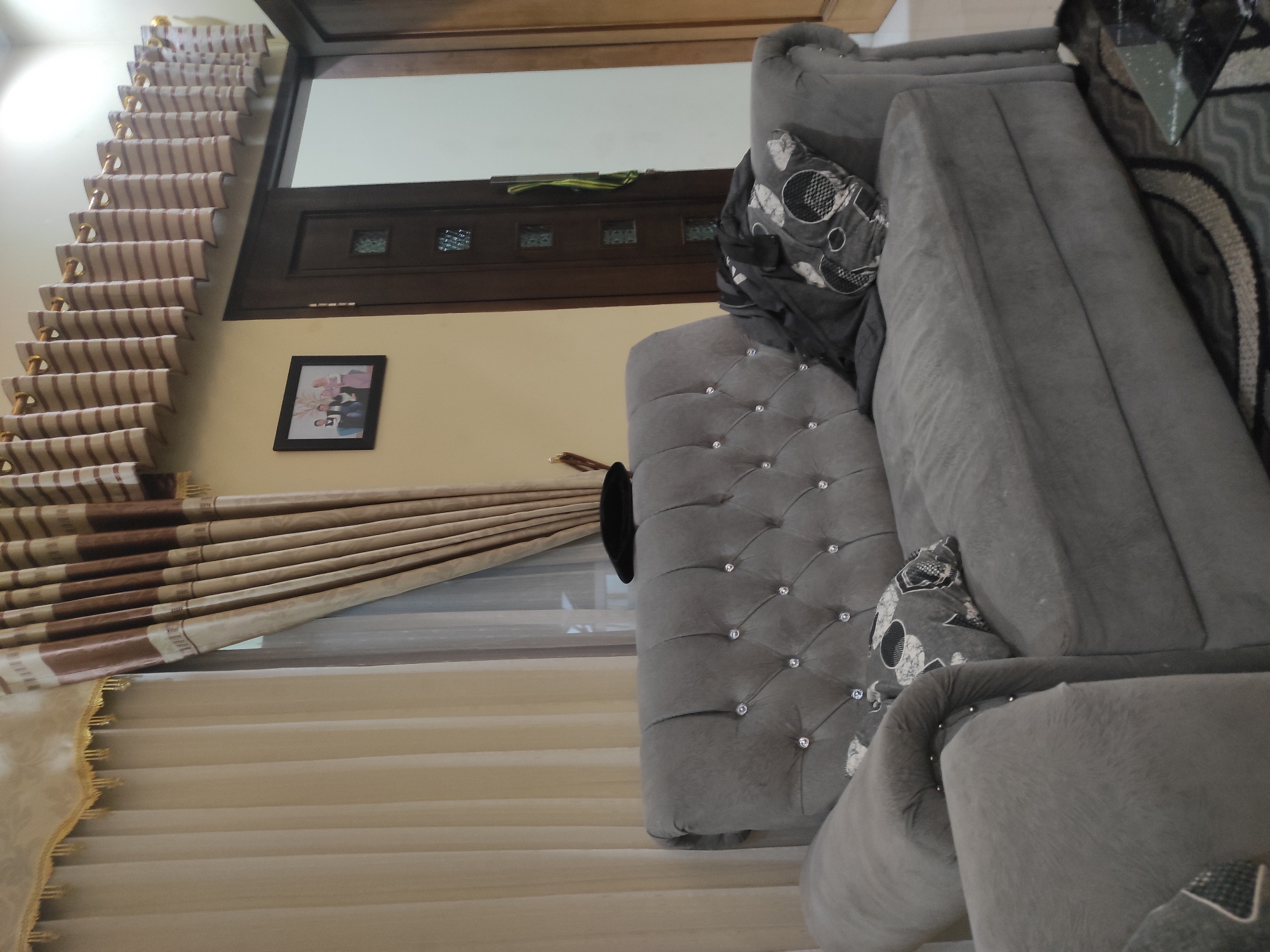 The image depicts an interior scene, likely a living room, in Sumedang, Indonesia. Dominating the foreground is a plush, grey, tufted sofa with diamond-like accents on the backrest. Two pillows are placed on the sofa, one a dark grey with a patterned design and another folded blanket or garment also in dark grey. To the left of the sofa, the armrest and a portion of another seating unit are visible. In the background, a wooden door with decorative glass panels is seen. Above the door, a set of horizontal curtains with brown and cream stripes hang, complemented by lighter cream curtains to the left. A framed photograph of four people, two adults and two children, is hung on the pale yellow wall above the door. The floor is partially covered by a rug with a geometric pattern. The lighting suggests an indoor evening setting. There are no visible individuals actively engaged in any activity.