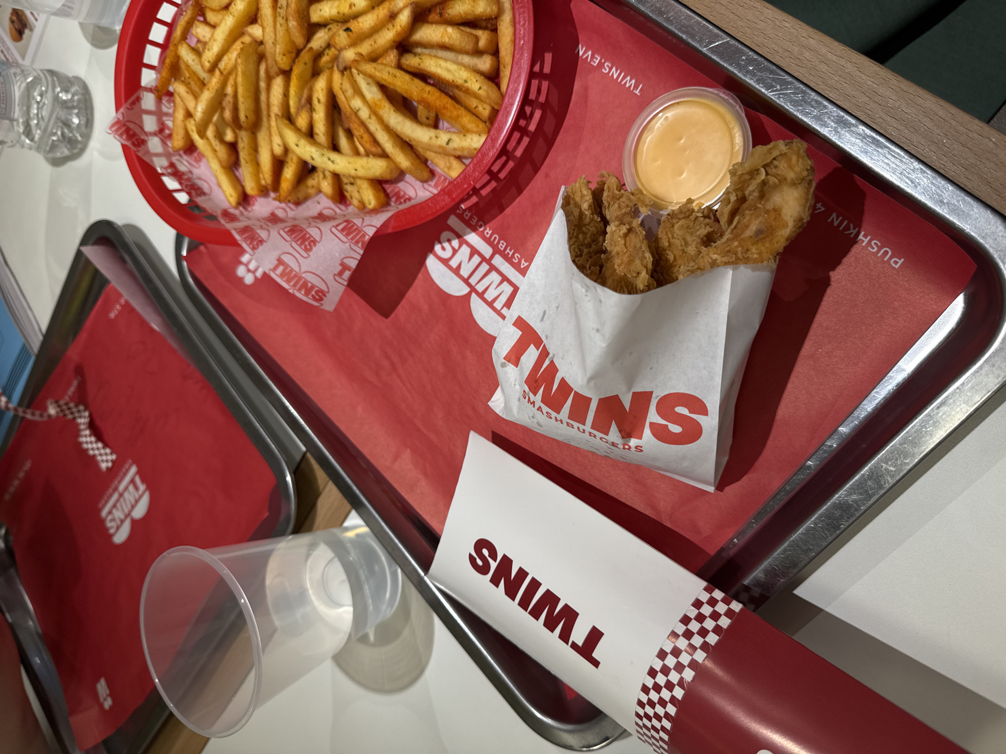 The image shows a meal from the restaurant "Twins Smashburgers" in Yerevan, Armenia. The meal is presented on a red branded tray with a metal frame. The food consists of a red plastic basket of french fries, a paper bag containing chicken strips, and a small container of dipping sauce. A plastic cup is nearby. The tray is placed on a table. The branding on the food packaging clearly identifies the restaurant.