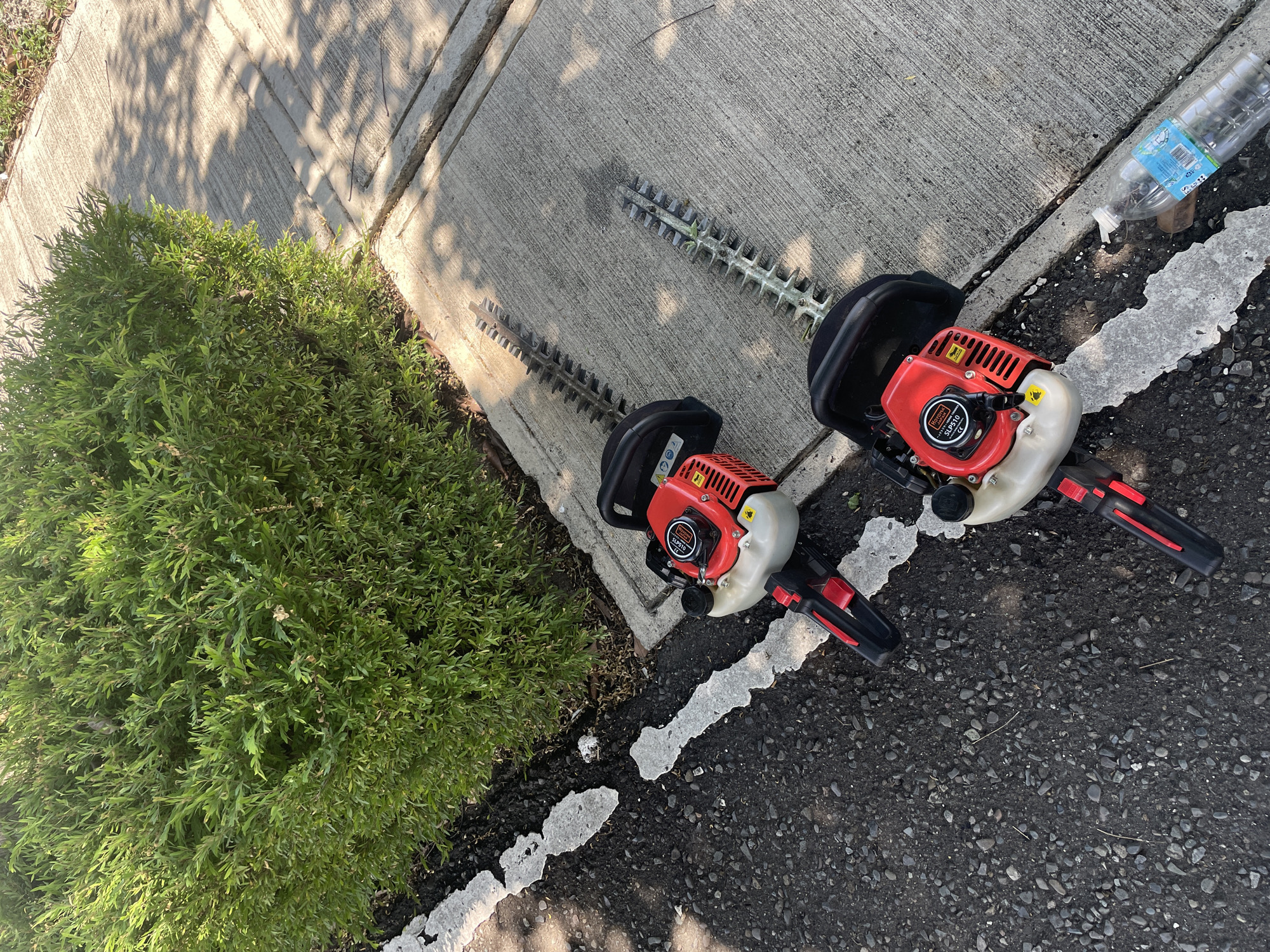 Two red and black gasoline-powered hedge trimmers are positioned on a concrete sidewalk next to a manicured green hedge. The hedge trimmers are angled diagonally across the frame, with their blades pointing towards the top left. One trimmer is positioned slightly behind and above the other. A clear plastic water bottle with a blue label is lying on its side on the sidewalk near the bottom right of the frame. The sidewalk is cracked in several places and is partially covered in shadows cast by sunlight filtering through unseen foliage. The area adjacent to the sidewalk is a dark asphalt surface with scattered small grey stones. The overall scene suggests outdoor maintenance or yard work in an urban or suburban setting within Davao City, Philippines, given the style of the hedge trimmers and the commonality of such tools for maintaining residential or public green spaces in the region.