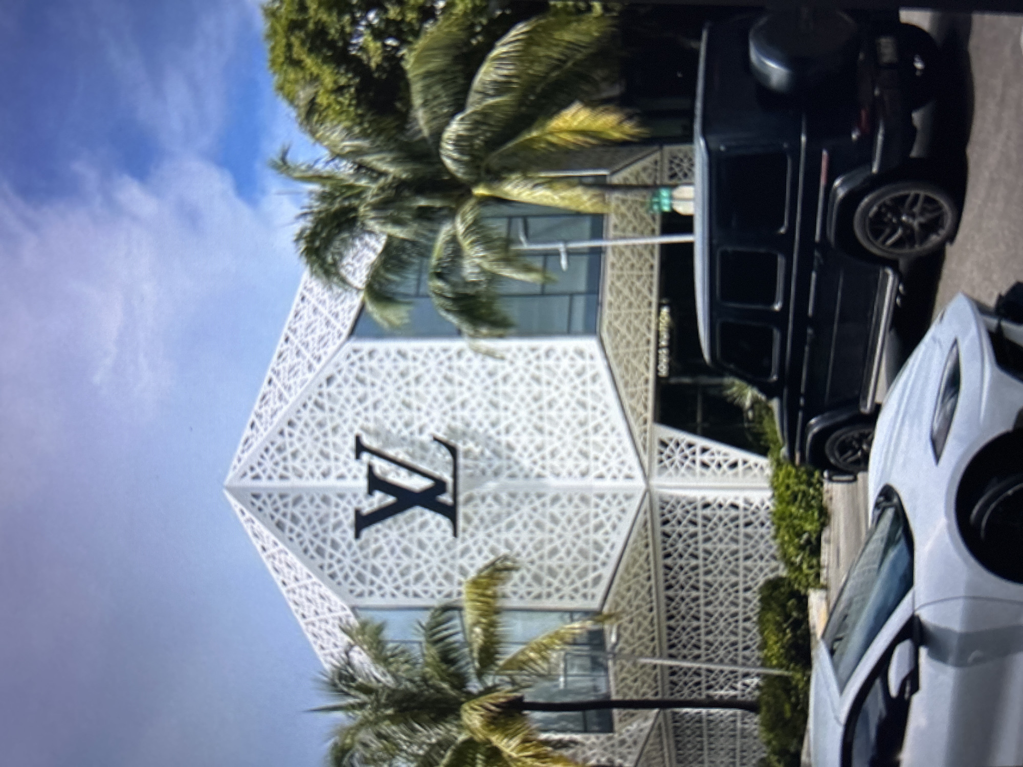 The image depicts the Louis Vuitton store, recognizable by its prominent LV logo, with a distinctive white geometric facade and tropical palm trees framing the scene. In the foreground, a white sports car is parked on the right, and to its right, a black SUV is visible. The sky above is blue with scattered clouds. The scene suggests a luxury shopping district in a warm climate, likely a city. However, there is no information available to confirm this is the city of Burgas, Bulgaria.
