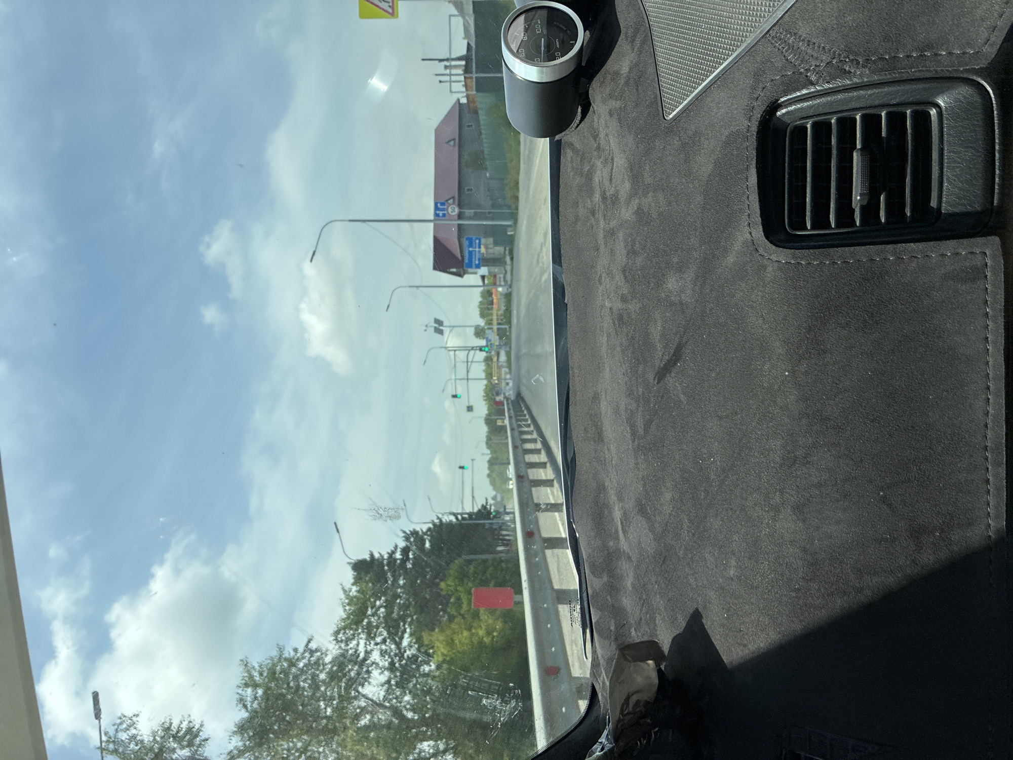 The image shows a view from inside a car, looking forward through the windshield. The dashboard is covered in a dark, textured material, with a black air vent visible in the lower right portion of the frame. A circular gauge with white markings and a needle is mounted on the dashboard on the right side. The road ahead is visible, with a metal guardrail on the left side. Trees line the left side of the road, and a red rectangular sign is visible among the trees. In the distance, there are buildings, traffic lights, and streetlights. The sky is blue with scattered white clouds. Based on the provided crops and information, the location is likely Spassk-Dal'nij, Russia, as suggested by contextual information.