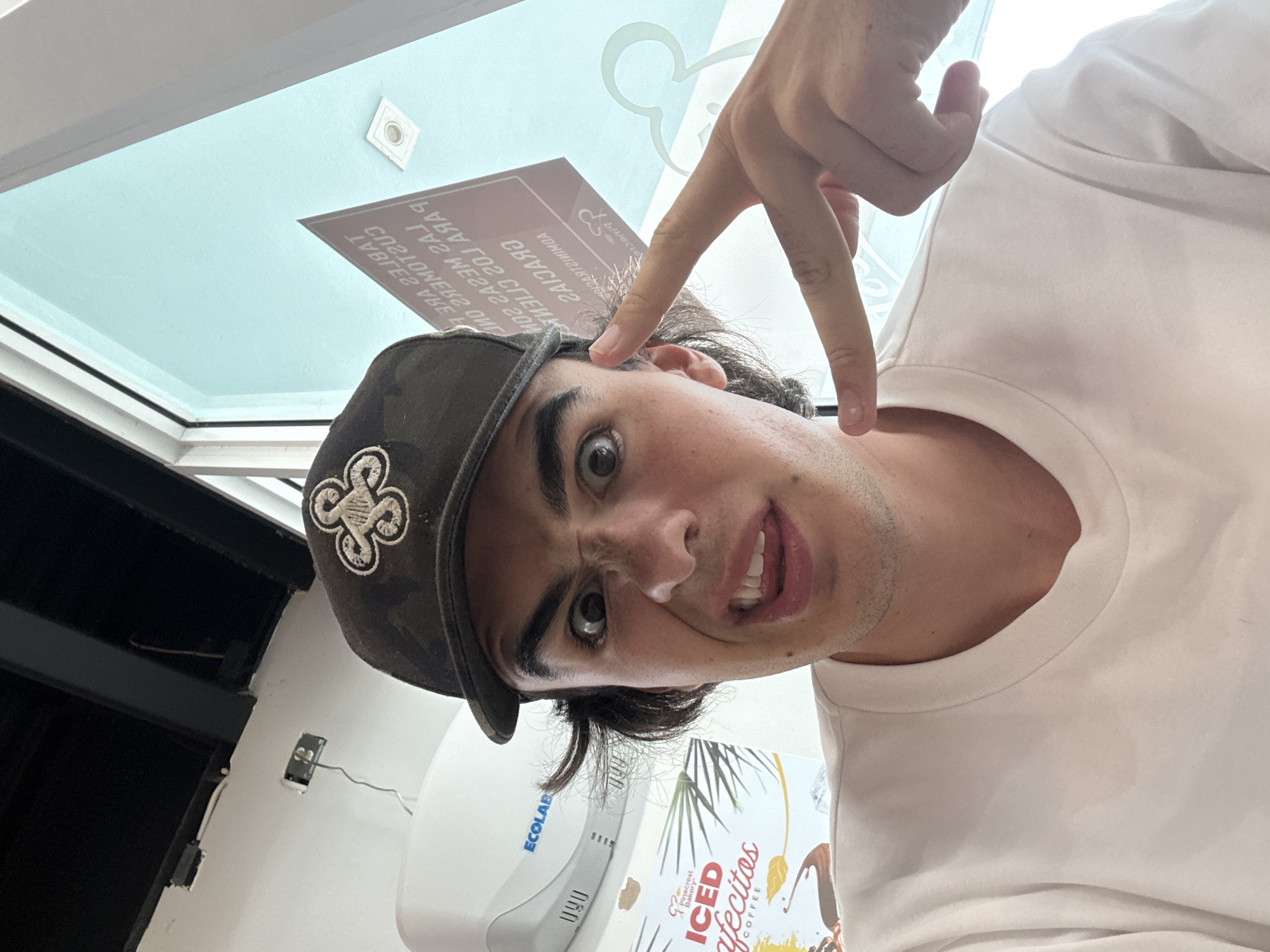 The image shows a selfie of a young man wearing a camouflage cap with a clover-like emblem, white t-shirt, inside a business. He's holding up his hand in a "peace sign" with the index finger pointed towards his temple. The business appears to be an establishment that serves "ICED Afecitos COFFEE," as seen on a sign. An Ecolab hand dryer is visible on the wall. The location is in Miami Beach, United States of America, as inferred from the text on the sign.