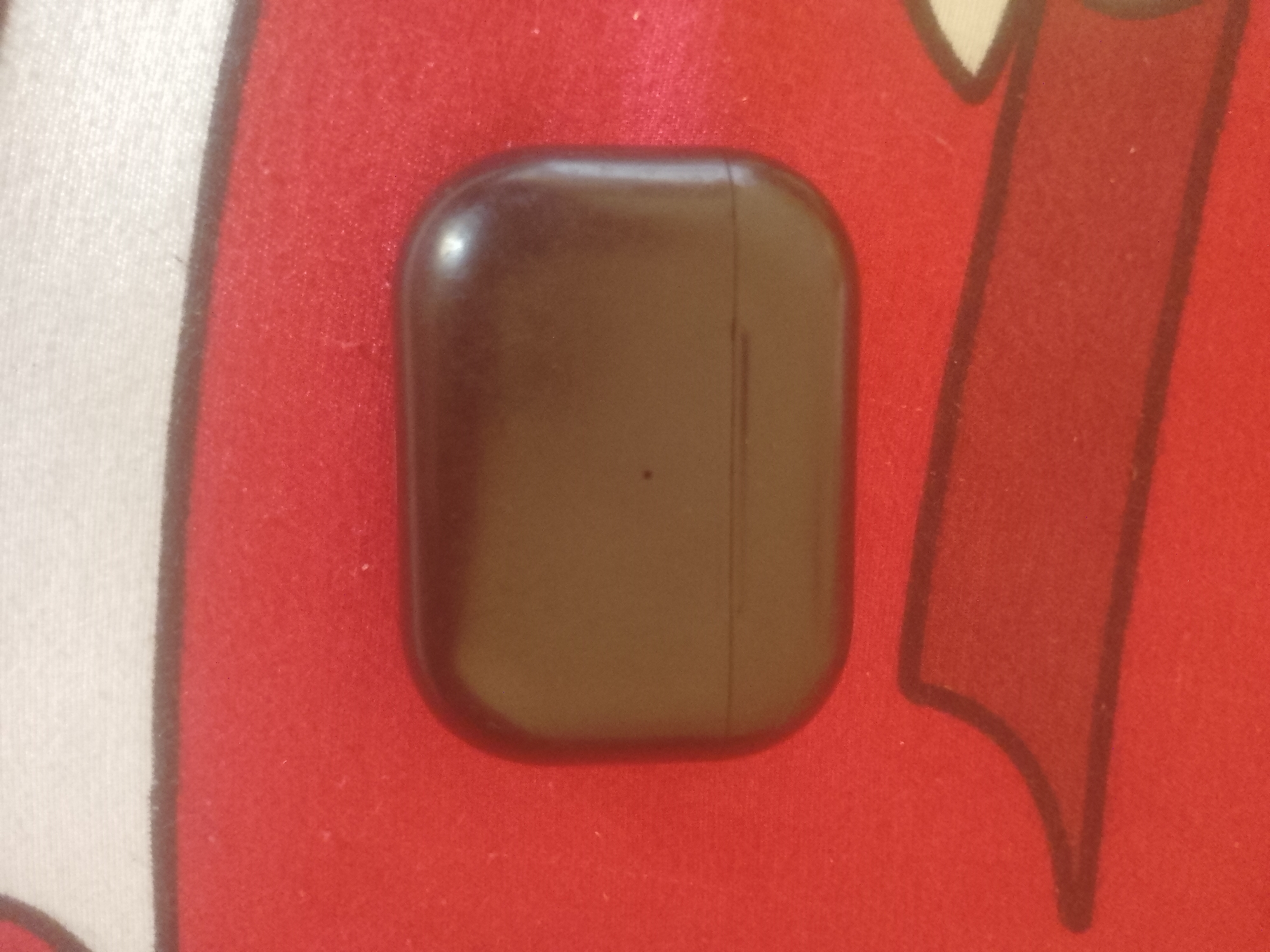 The image shows a brown, rectangular object with rounded corners resting on a red surface. The object appears to be a charging case for earbuds. The red surface has a textured pattern, possibly fabric, with some white and black elements visible in the background, suggesting a patterned mat or tablecloth. The lighting is somewhat dim, casting a slight shadow. There is a faint vertical line down the center of the object, indicating a seam. There are no people or animals visible in the image, nor any specific urban or natural landmarks that would indicate a location in Majemu, Nigeria. The image is a close-up of the object and its immediate surroundings.