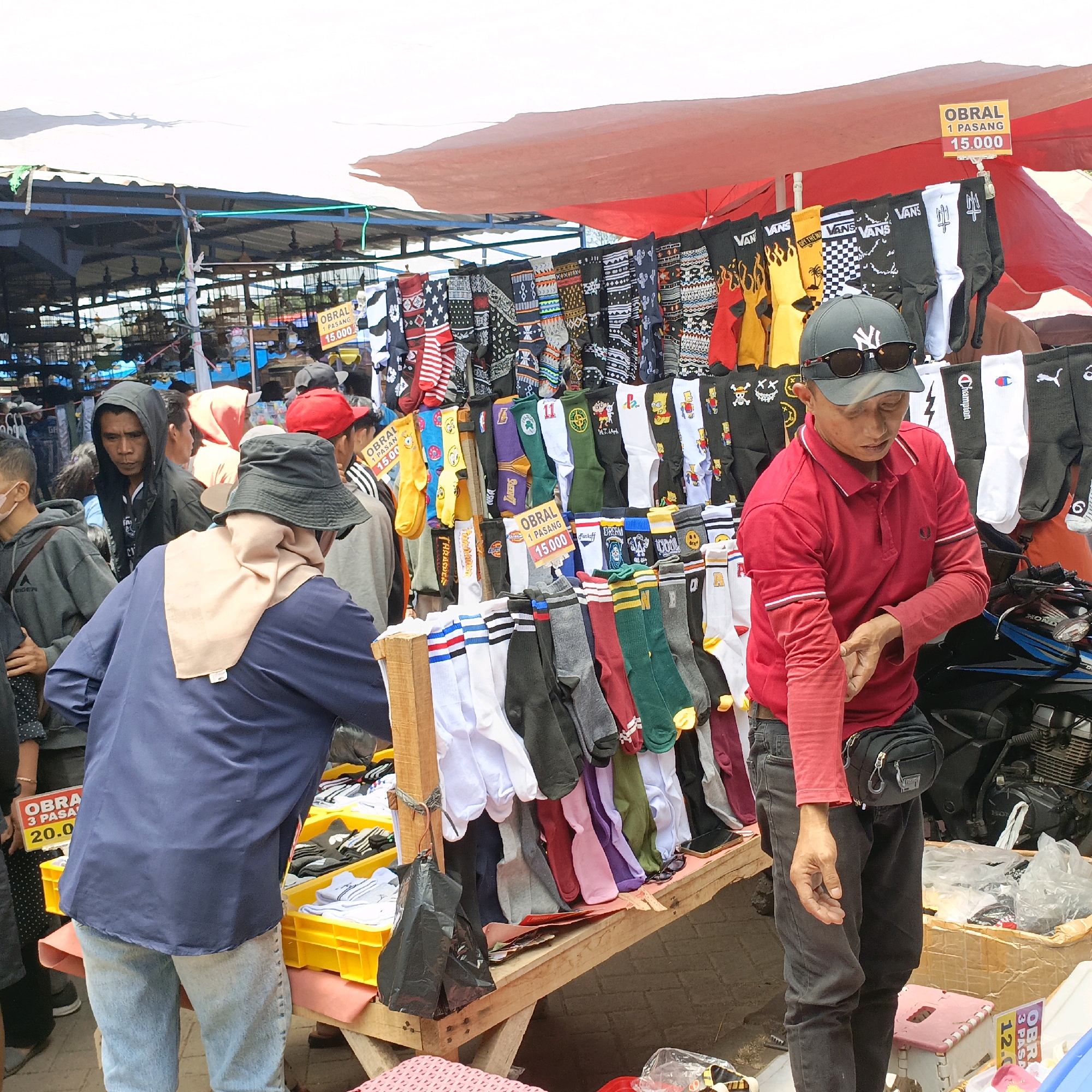 The image captures an outdoor market scene, likely in Ambarawa, Indonesia, given the context. A stall is set up with a variety of socks displayed for sale, featuring diverse patterns and colors, including those with cartoon characters like Bart Simpson and designs with logos like Vans and Champion. The socks are hung on a rack and also displayed in yellow plastic crates. Prices are indicated on signs, with one reading "OBRAL 1 PASANG 15.000".
Several people are present around the stall. A man in a red polo shirt and dark pants, wearing a New York Yankees baseball cap and sunglasses, appears to be attending the stall, adjusting his sleeve. Other individuals, some wearing hoodies and casual clothing, are browsing or standing near the stall. One person is wearing a blue long-sleeved shirt and a beige headscarf, while another is seen in a grey hoodie. A motorcycle is partially visible in the background, suggesting a typical street market environment. The stall itself is covered by a reddish-pink tarp. The ground appears to be paved with bricks. The overall atmosphere suggests a busy commercial area.