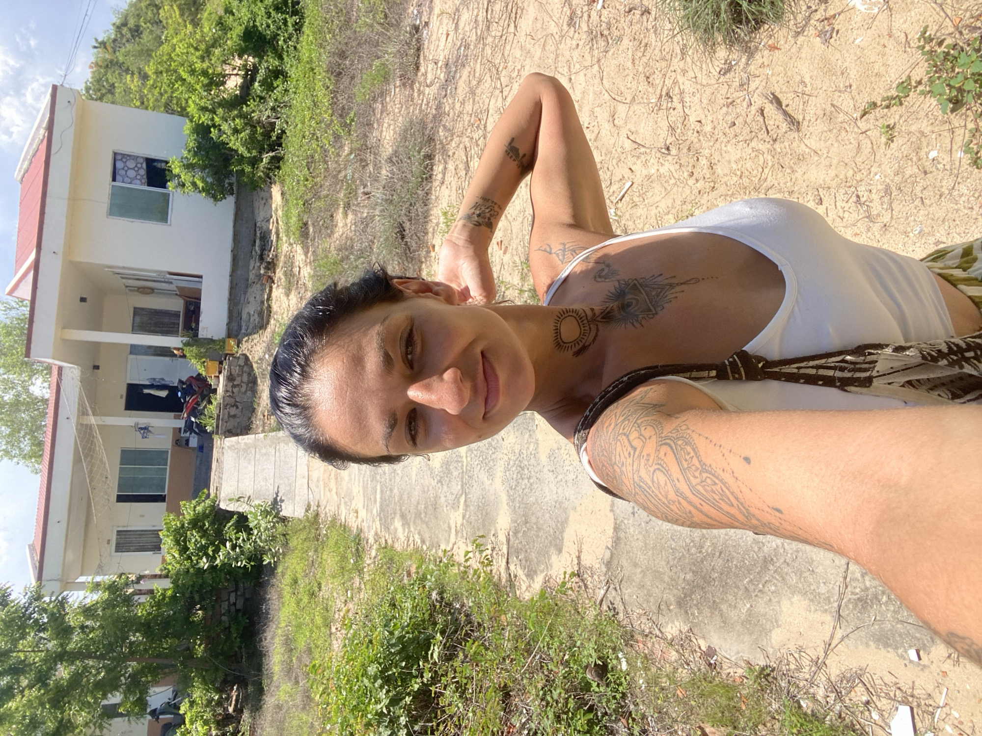 A selfie is taken by a woman with dark hair and several tattoos, standing outside a white house with a red roof in Mũi Né, Vietnam. She is wearing a white tank top and a patterned skirt. The house has a concrete walkway leading up to it, and is surrounded by vegetation and sandy ground. There are motorcycles parked in front of the house. The sky is cloudy.