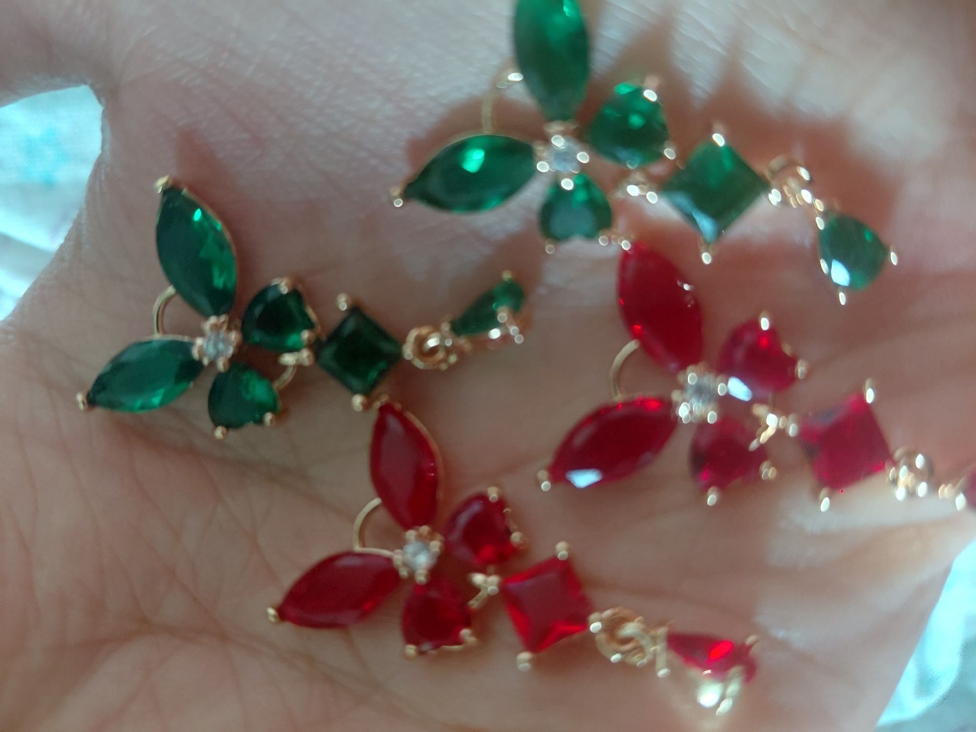 The image displays six individual pieces of jewelry, possibly earrings or charms, arranged on the open palm of a light-skinned human hand. Each piece features gold-toned metal settings encasing various cut gemstones. The gemstones are predominantly deep red, appearing as marquise and square cuts, and emerald green, appearing as marquise, square, and teardrop cuts.
Each primary jewelry cluster is designed as a four-petal flower or butterfly, with four marquise-cut stones (either red or green) radiating from a central small, round, clear gemstone. Attached to some of these clusters are dangling elements, typically consisting of a single square-cut stone followed by a smaller teardrop-cut stone, matching the color scheme of the main cluster (either red or green).
The hand, with visible creases and skin texture, serves as the foreground. The background is unfocused, suggesting soft, light-colored fabric, possibly light blue in tint, behind the hand.