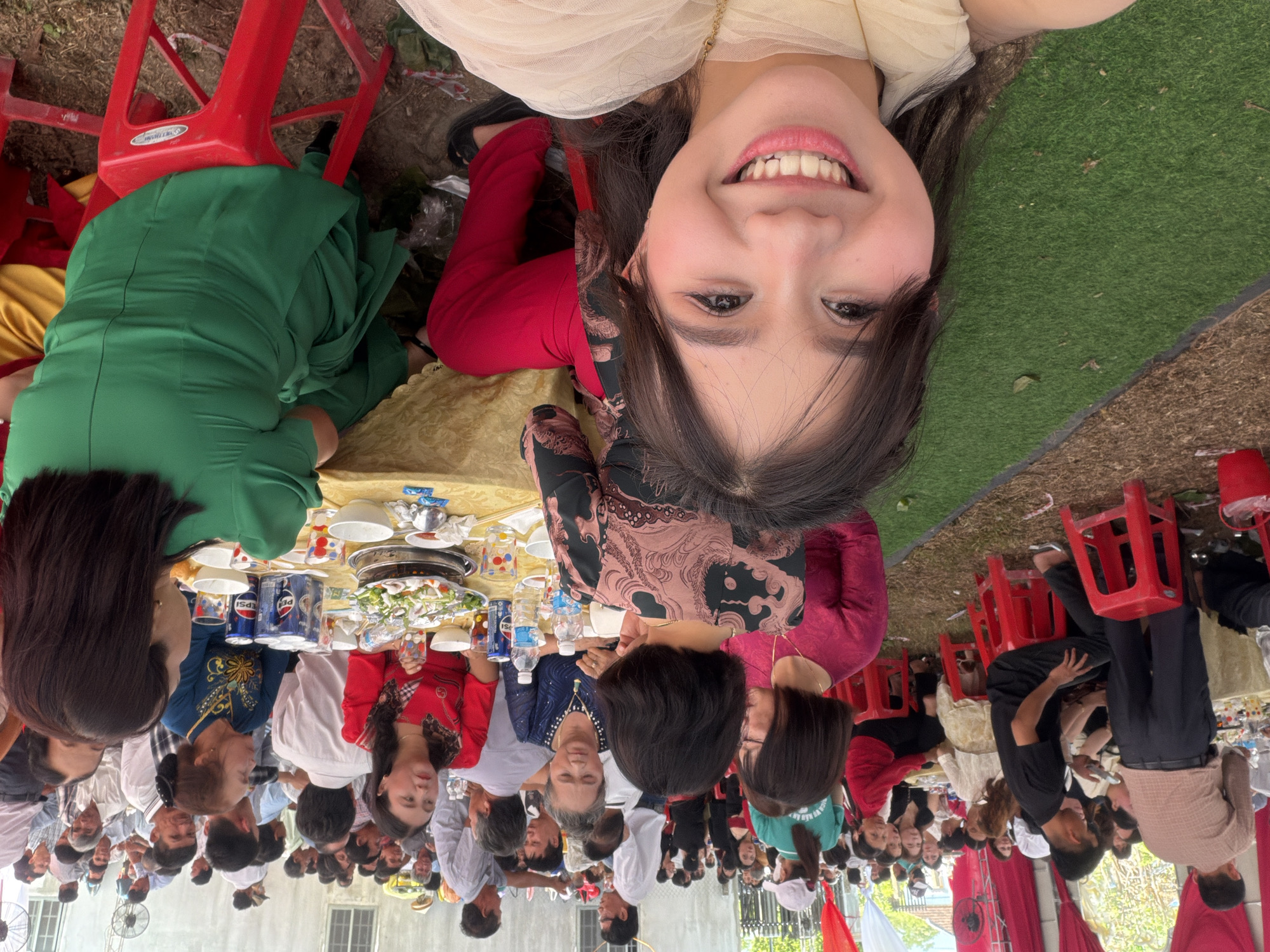 The image, captured from a first-person perspective, is rotated counter-clockwise, showcasing a lively social gathering in Tay Son Commune, Vietnam. In the immediate foreground, a young woman with dark hair and a radiant smile looks directly at the viewer. She is wearing a cream-colored sheer top, red pants, and shoes with intricate patterns, conveying a sense of happiness and engagement.
Behind her, numerous individuals are seated around multiple tables, participating in a communal meal. The tables are covered with golden yellow tablecloths and abundant with food, including a large central dish, likely a hot pot, along with various plates, bowls, and beverages. Cans and bottles of "PEPSI" are clearly visible on the tables, alongside bottled water. The attendees, mostly adults dressed in a mix of casual and semi-formal clothing, are seen eating and interacting. Some women wear traditional-style tops.
The gathering appears to be held outdoors or under a large, open-sided structure, with patches of green artificial turf and bare earth visible on the ground. Bright red plastic chairs are a prominent feature throughout the scene. In the background, simple buildings and a large standing fan suggest a warm, daytime environment. The overall atmosphere is one of conviviality and celebration.
