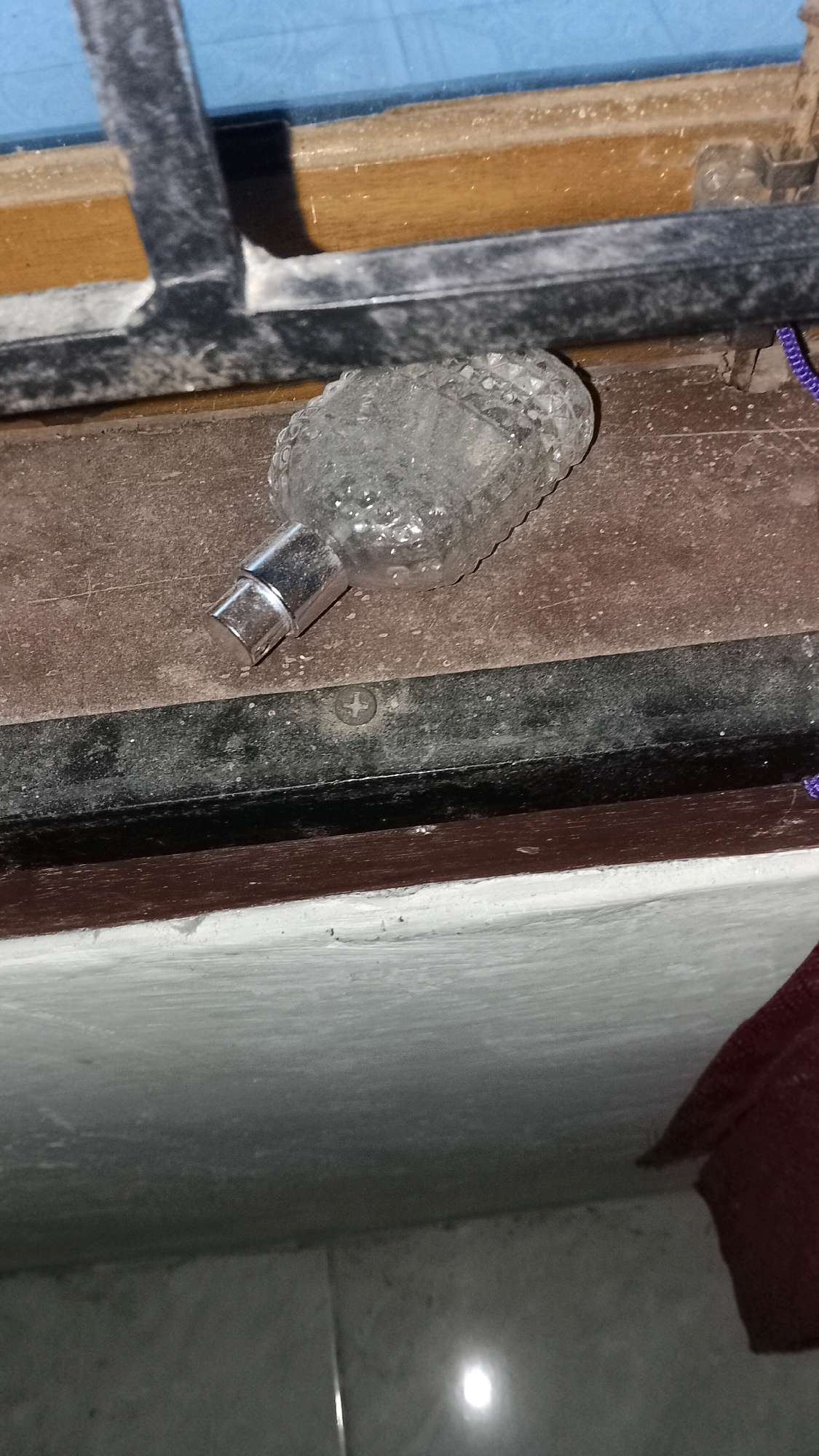 A clear, empty perfume bottle with a silver cap lies on a dusty, dark brown surface. The bottle is textured and appears to be made of glass. To the left, a dark metal window frame is visible, with a wooden structure behind it and a blue surface above. Below the brown surface is a black metal strip with a visible screw. Further down, a white ledge is present, and the floor below is tiled and reflects a bright light source. The scene is indoors, likely in a domestic setting, given the presence of a window frame and floor tiles. The location is Mulyo Haji, Indonesia.