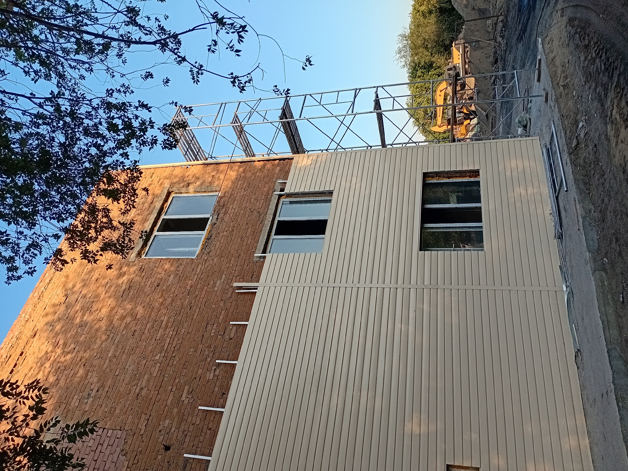 The image depicts the exterior of a brick building undergoing renovation, located in the city of Semey, Kazakhstan. The lower section of the building is clad in beige siding, while the upper section remains exposed brick. Scaffolding is erected on the right side of the building, suggesting ongoing construction work. A clear blue sky is visible in the background, with branches of a tree casting shadows on the building. In the distance, construction equipment, including a yellow excavator, is present on a dirt ground. The foreground shows a concrete walkway with metal pipes and construction debris.