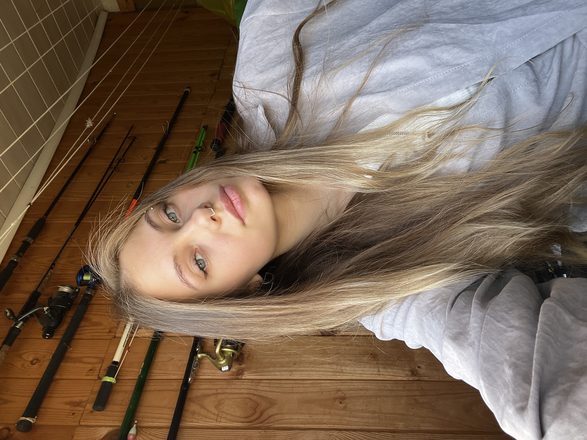 A young woman with long blonde hair is lying on a wooden surface, possibly a deck or floor, indoors. She wears a light grey hooded jacket. Several fishing rods and reels are visible along the left side of the image. She has a nose ring and possibly an eyebrow piercing. The location is not evident in the image. It is not possible to determine the location is Фоки, Russia.