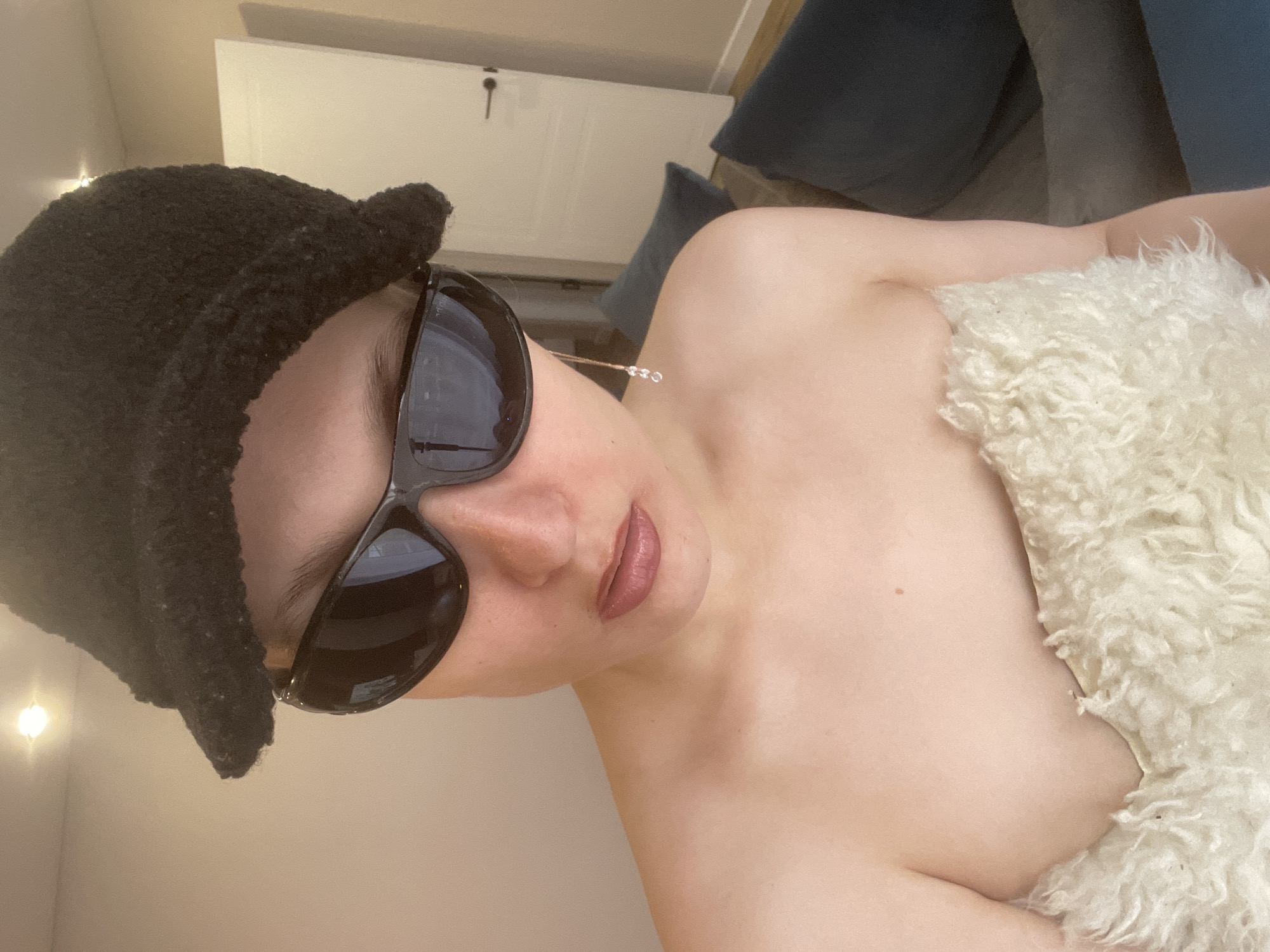 A woman wearing sunglasses and a dark beanie is lying down. A white, fluffy material is draped over her body. The location is not identifiable as Подшивалово, Russia based on the provided image.