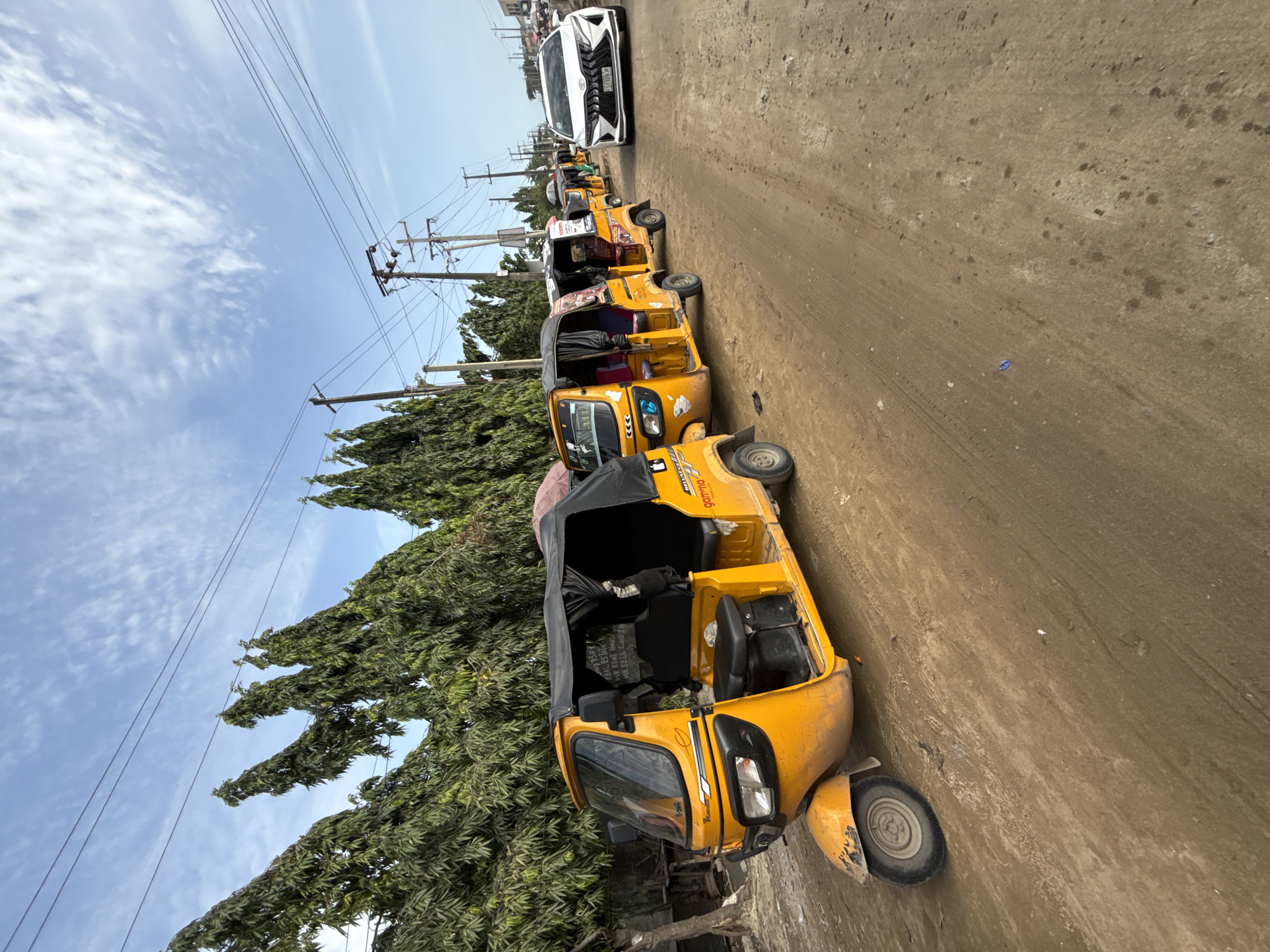 The image depicts a line of yellow "keke napep" auto rickshaws parked on a dirt road in Lekki, Nigeria. A white vehicle is parked further down the road. Power lines are visible overhead, and trees line the side of the road. The sky is blue with some clouds.