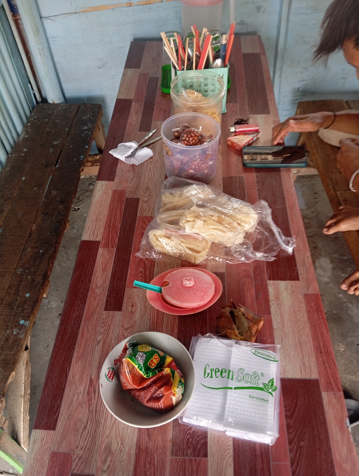 A high-angle medium shot captures a wooden table laid with food and utensils in what appears to be an outdoor eating area. The table's surface is a patterned laminate with various shades of brown and red, resembling wood grain. To the left, a rough, wooden bench with visible wear and tear is partially visible, suggesting it's part of the seating arrangement.
On the table, several items are arranged. In the background, a collection of chopsticks and spoons are held in a green container. Next to it, a clear plastic tub holds a brown, granular substance. Closer to the foreground, a clear plastic bag contains flat, light-colored food items, possibly crackers or chips. A pink plate holds a round, pink object, perhaps a type of dessert or frozen treat, with a teal utensil stuck into it. A small, brown object, resembling a dried food item or shell, sits next to the plate. To the right, a white packet of "Green Soft" napkins is prominently displayed. A white bowl contains a crinkled, colorful plastic wrapper.
In the mid-right of the frame, a person's hands are visible, reaching towards a cigarette lighter and a small, dark rectangular object, possibly a phone or a wallet, resting on the table. The person's arms and part of their head are also visible, indicating they are seated at the table. Their right hand appears to be wearing a bracelet. Their bare feet are visible at the bottom right of the image, suggesting a casual setting. The background shows a wall constructed of vertical planks and some metallic siding. The overall impression is a simple, informal setting where food is being served or consumed. The presence of local brands like "Green Soft" and the general aesthetic suggest a location in a town or village in Indonesia, potentially Sokaraja Wetan.