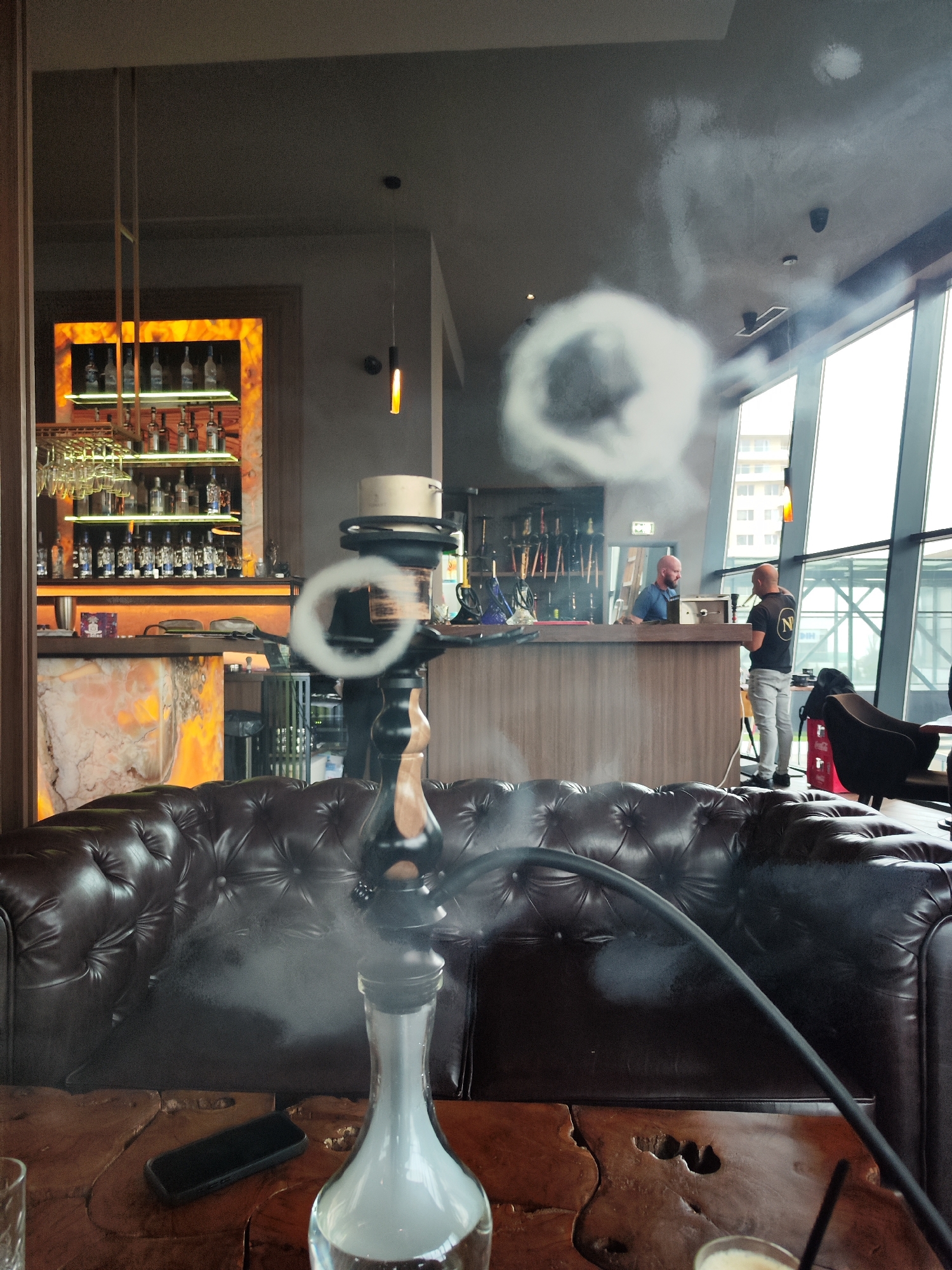 This image depicts the interior of a modern bar or hookah lounge, likely in Burgas, Bulgaria. In the foreground, a stylish black and wood hookah with a clear glass base containing milky white smoke is prominently featured on a rustic wooden table. A black hose extends from the hookah, and several distinct smoke rings are visible in the air, including one directly in front of the hookah's stem and a larger one drifting towards the ceiling. A black phone case and a glass are also on the table.
Behind the foreground elements, a large, dark brown tufted leather sofa provides comfortable seating, suggesting a relaxed lounge atmosphere.
In the mid-ground, a contemporary bar area is visible. The bar counter features an illuminated backsplash made of what appears to be marble or onyx, showcasing rows of various liquor bottles and suspended wine glasses. Two bald men are behind the counter; one in a blue shirt is further back, while the other, wearing a black t-shirt with a white stylized 'N' logo within a circle, stands closer to the right. They appear to be conversing or attending to tasks at the bar.
The background features large windows offering a view of an urban environment. A multi-story building with numerous windows is visible under an overcast, grey sky, suggesting either a cloudy day or late afternoon/early evening. Interior lighting, including a modern pendant light with an orange glow and recessed spotlights, is active. The ceiling shows evidence of smoke (from the hookah), smoke detectors, and additional lighting fixtures. The overall scene conveys a relaxed and social ambiance within a chic, contemporary establishment.