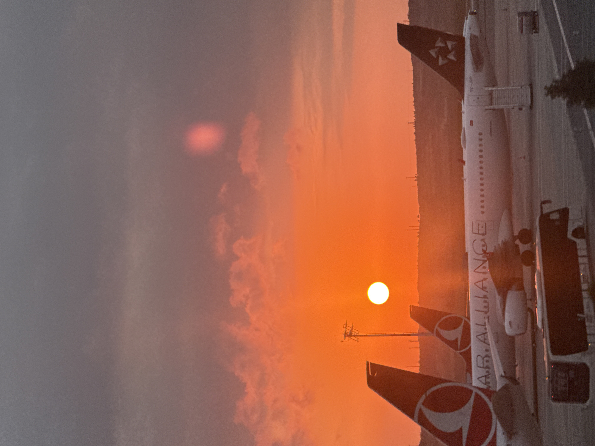The image shows an airport scene at sunset in Arnavutköy, Turkey. Three airplanes are parked on the tarmac. The sky is a vibrant orange and red, with some clouds. The sun is setting, casting a warm glow over the scene. A bus is parked in the foreground, and a luggage cart is visible near one of the airplanes. The overall impression is one of a peaceful and serene evening at the airport.