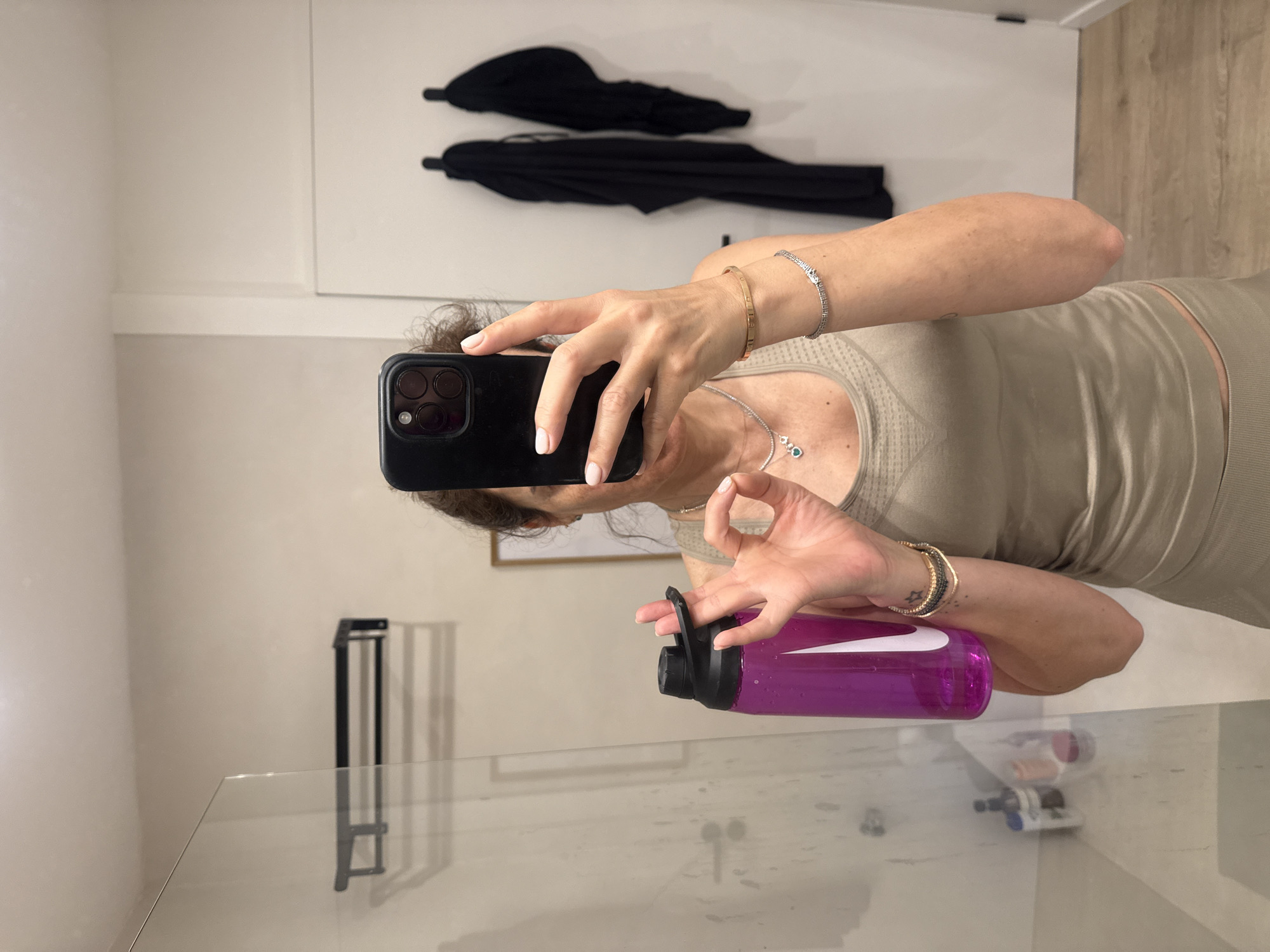 An indoor mirror selfie shows a woman, partially obscured by her smartphone, capturing her reflection. The image is oriented sideways, with the wall appearing on the left and upper sections of the frame.
The woman, with brown hair, is wearing activewear consisting of a light, possibly beige or olive green, ribbed tank top and matching bottoms. She holds a black smartphone, likely an iPhone, in her left hand, which covers most of her face. Her nails are painted a light, neutral color.
Her left wrist displays two bracelets: one appears to be a gold-toned bangle, and the other a silver-toned bracelet adorned with small stones. Around her neck, she wears a silver necklace with a pendant featuring a prominent green stone surrounded by smaller clear gems. In her right hand, she holds a translucent purple water bottle with a black lid and a visible white "Nike" logo. On this wrist, she also wears a gold-toned bracelet and a silver-toned bracelet with stones, similar to the one on her left wrist.
The background behind her features a light-colored wall with two black garments or umbrellas hanging on hooks. To the right, a wooden panel or flooring is visible. The lower part of the frame reveals a clear glass surface, possibly a table or counter, reflecting elements of the room and some objects beneath it, including a black metal structure. The setting appears to be a well-lit interior, possibly a home or gym, in Vila Nova de Gaia, Portugal. The overall activity is a casual self-portrait.