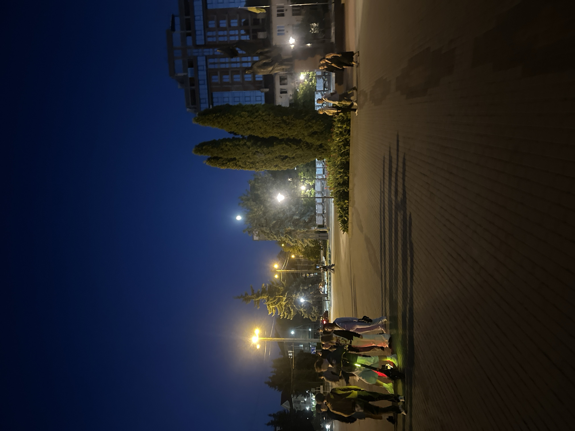 This is a nighttime photograph taken in Ternopil, Ukraine, likely in a public square. In the foreground, a group of young people are gathered, some standing and some seemingly preparing to ride an electric scooter that is illuminated with green and red lights. The pavement is paved with rectangular blocks, and long shadows are cast by the group and streetlights.
In the midground, several tall streetlights illuminate the scene with warm yellow light. Behind the young people, more trees are visible, including some large, mature evergreen trees. Further back, a modern building with glass windows dominates the skyline, reflecting the night sky. To the right of this building, a statue is visible in front of a smaller, older building.
The sky is a deep, dark blue, with a bright full moon visible in the upper center of the frame. Other lights from buildings and streetlights are present in the background, suggesting an urban environment. The overall atmosphere is calm and social, with individuals and small groups present in the square.