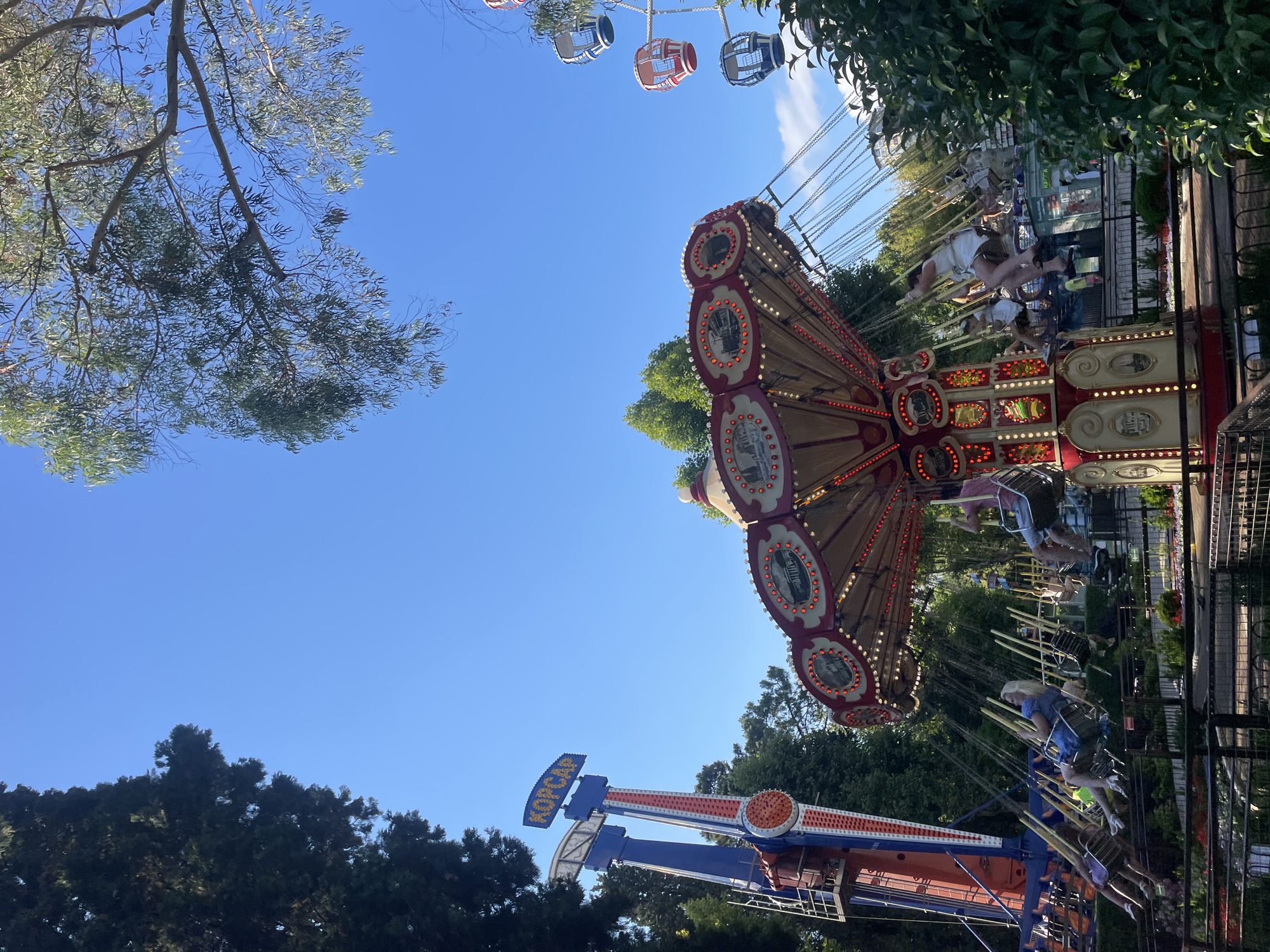This is a photograph taken at an amusement park in Sochi, Russia, likely during daylight hours given the bright sky and illuminated rides. The image features a swing ride in the foreground, with its red and gold canopy decorated with lights and circular scenes. Several people are seated on the swings, which are suspended by chains and appear to be in motion. To the left of the swing ride is a tall, blue and red amusement park ride with the word "КОРСАР" (KORSAR) visible at the top. Further in the background, to the right, a Ferris wheel with red and blue gondolas can be seen against the clear blue sky. Lush green trees surround the amusement park attractions, suggesting an outdoor, park-like setting. The overall scene depicts a vibrant amusement park experience with active rides and people enjoying themselves.