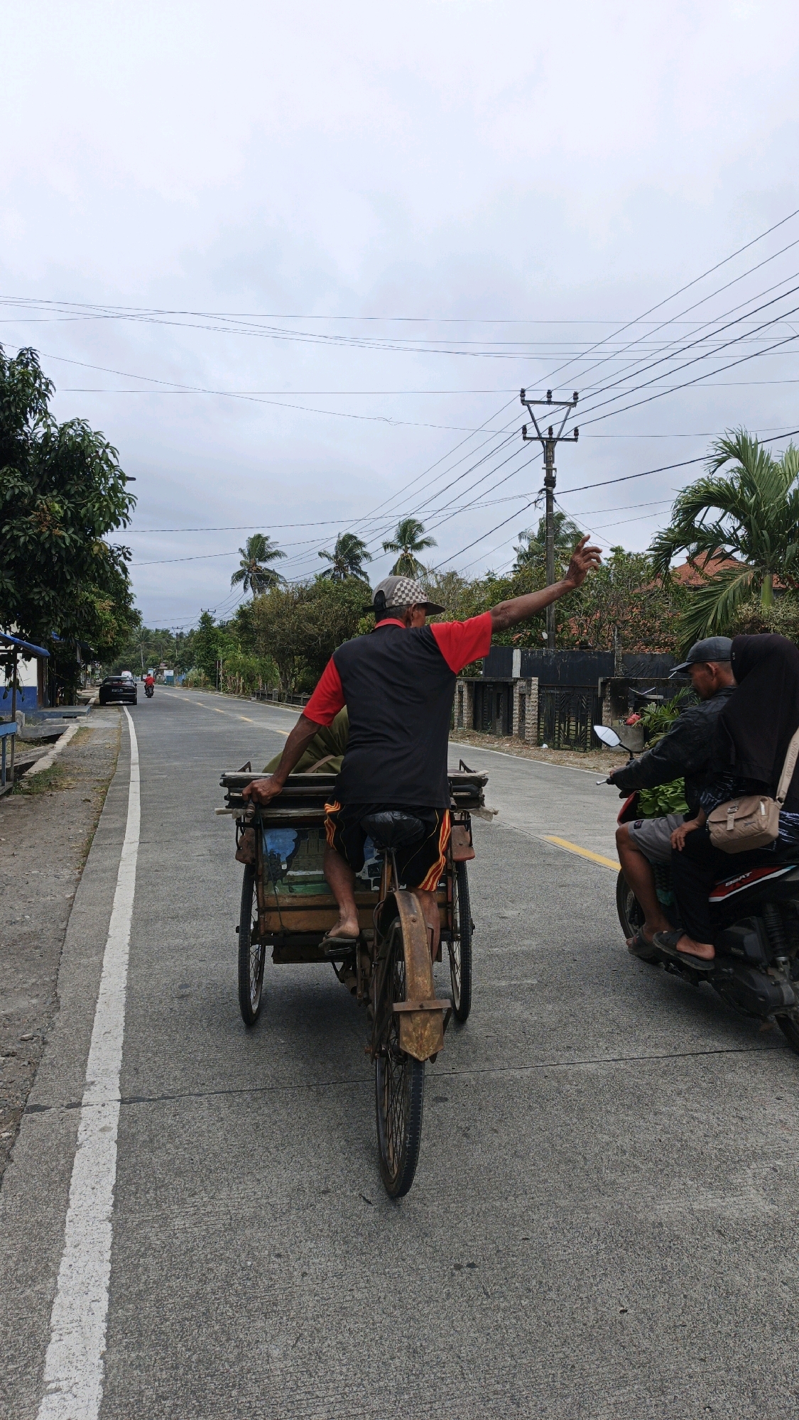 An overcast, daytime, eye-level medium shot captures a man riding a pedal-powered tricycle, which is hauling a cargo box, down a paved road in Cibenda, Indonesia. The man, facing away from the camera, wears a checkered baseball cap, a black t-shirt with red sleeves, and dark shorts. He has his left arm raised, palm open, in a wave or gesture. Behind him, a motorcycle is visible, carrying two passengers. The road is marked with a white line and stretches into the distance, lined with trees, palm trees, and buildings. Power lines and a utility pole are also present in the scene. The overall environment suggests a rural or semi-urban setting.