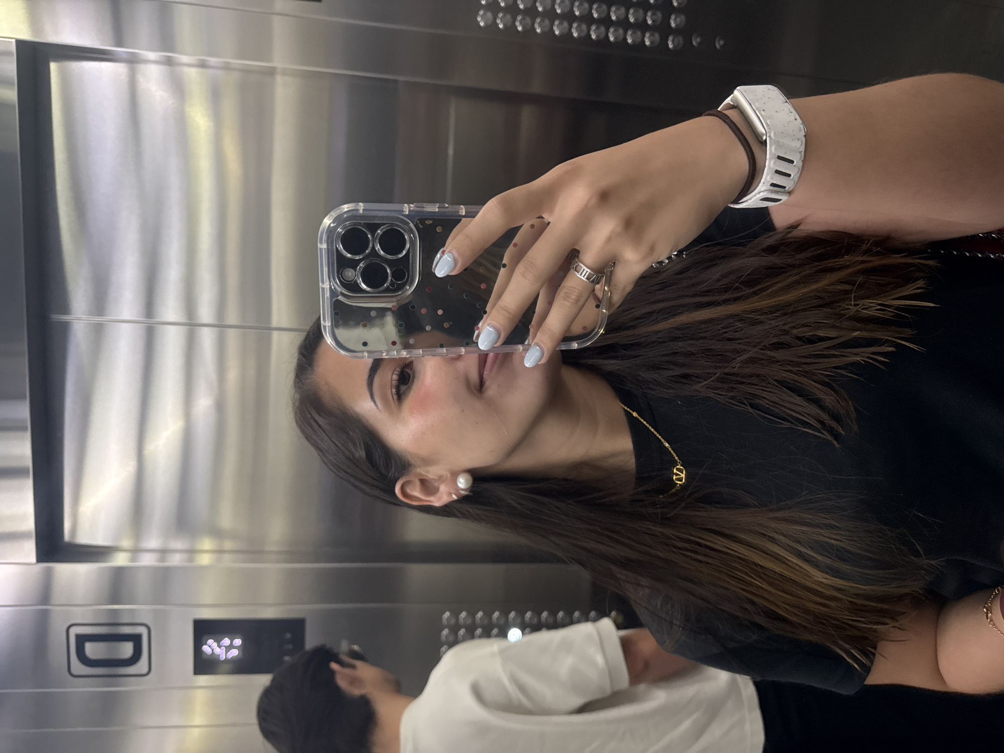 A woman is taking a selfie in an elevator. She is holding a smartphone with a clear case that has colorful polka dots, with light blue nail polish. She is wearing a white smart watch on her left wrist, a silver ring on her left ring finger, and a gold necklace with a "V" charm. She has brown hair and a pearl earring.
Behind her, another person wearing a white shirt is visible. The elevator has a brushed metal interior, with buttons and a "D" symbol visible. The location is unknown and there is no information indicating this is San Bernardino Tlaxcalancingo, Mexico.