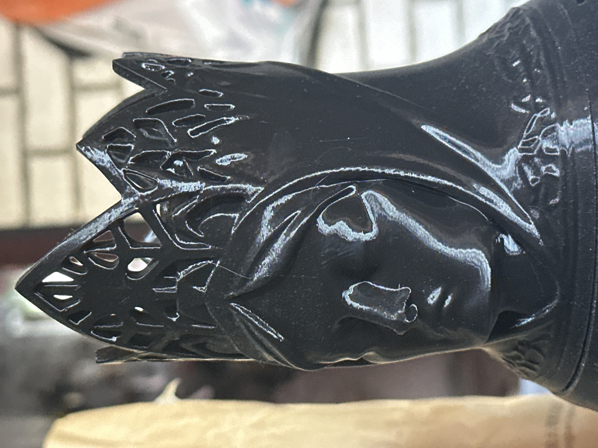 A close-up photograph depicts a black, possibly 3D-printed, object with intricate lattice work at one end and a smoother, rounded body. The object's surface appears glossy with visible print lines and some imperfections. It rests on a light brown surface. The image has a shallow depth of field. There is no information to indicate that this image was taken in Zelenograd, Russia.