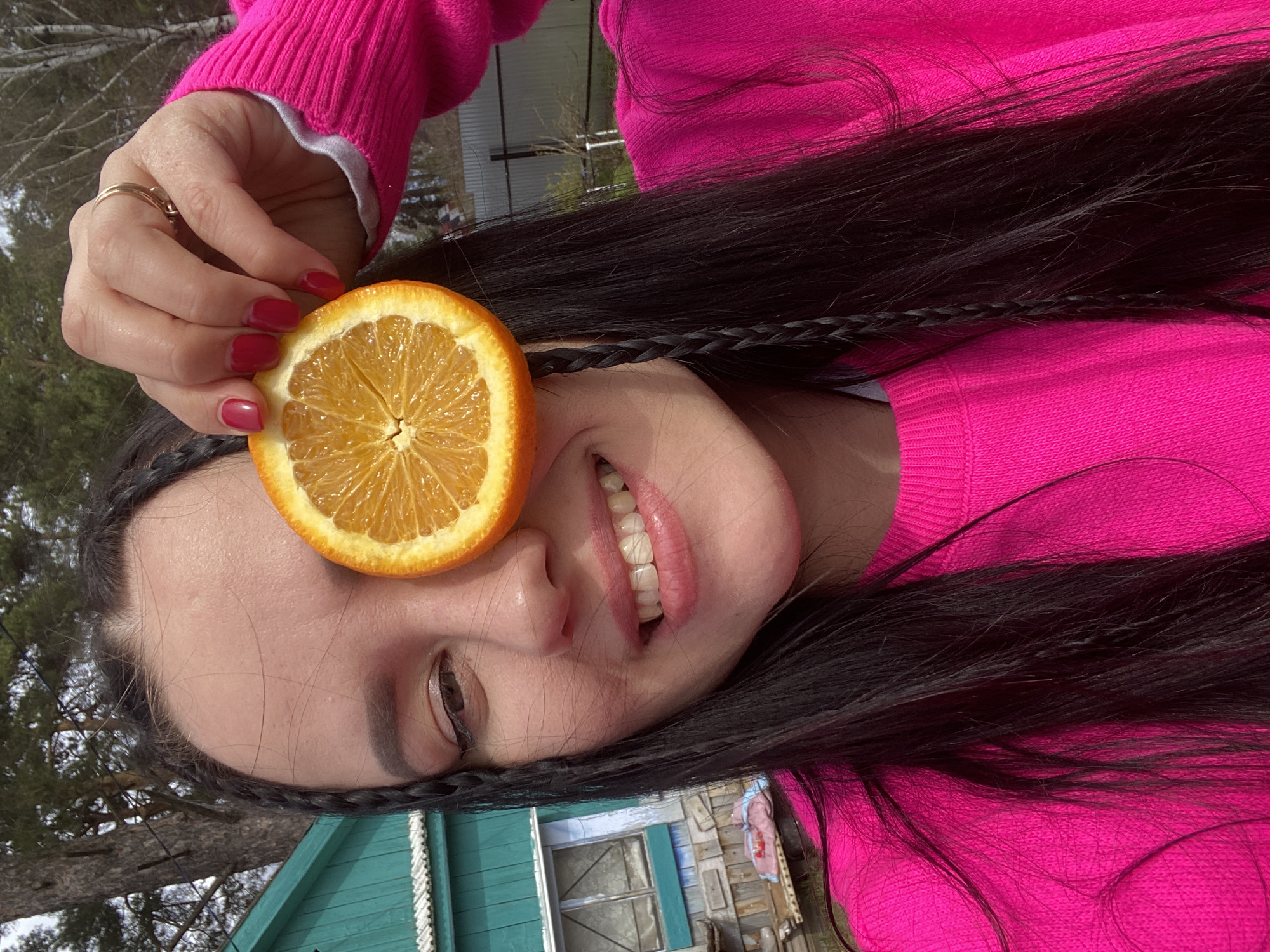 A vibrant outdoor portrait features a young woman posing playfully, with the image rotated 90 degrees counter-clockwise. The woman, shown from the upper chest upwards, has long, dark hair styled with at least one thin braid near her forehead. She is wearing a bright fuchsia-pink long-sleeved sweater. Her fingernails are painted a reddish-pink, and she wears a gold ring on one finger.
She holds a sliced half of an orange over her left eye, obscuring it. The woman has a pleasant, gentle smile, showing her teeth, and her right eye is open, looking towards the viewer. Her expression conveys a lighthearted and cheerful mood.
The background indicates an outdoor setting, likely a rural or semi-rural area, consistent with the provided location context of Chaikovsky, Russia. Bare trees or trees with sparse foliage are visible against a bright, natural light, suggesting daytime and potentially early spring or late autumn. A distinct teal or greenish wooden building with a window and siding is partially visible in the background, adding to the rustic aesthetic. No text is visible in the image.