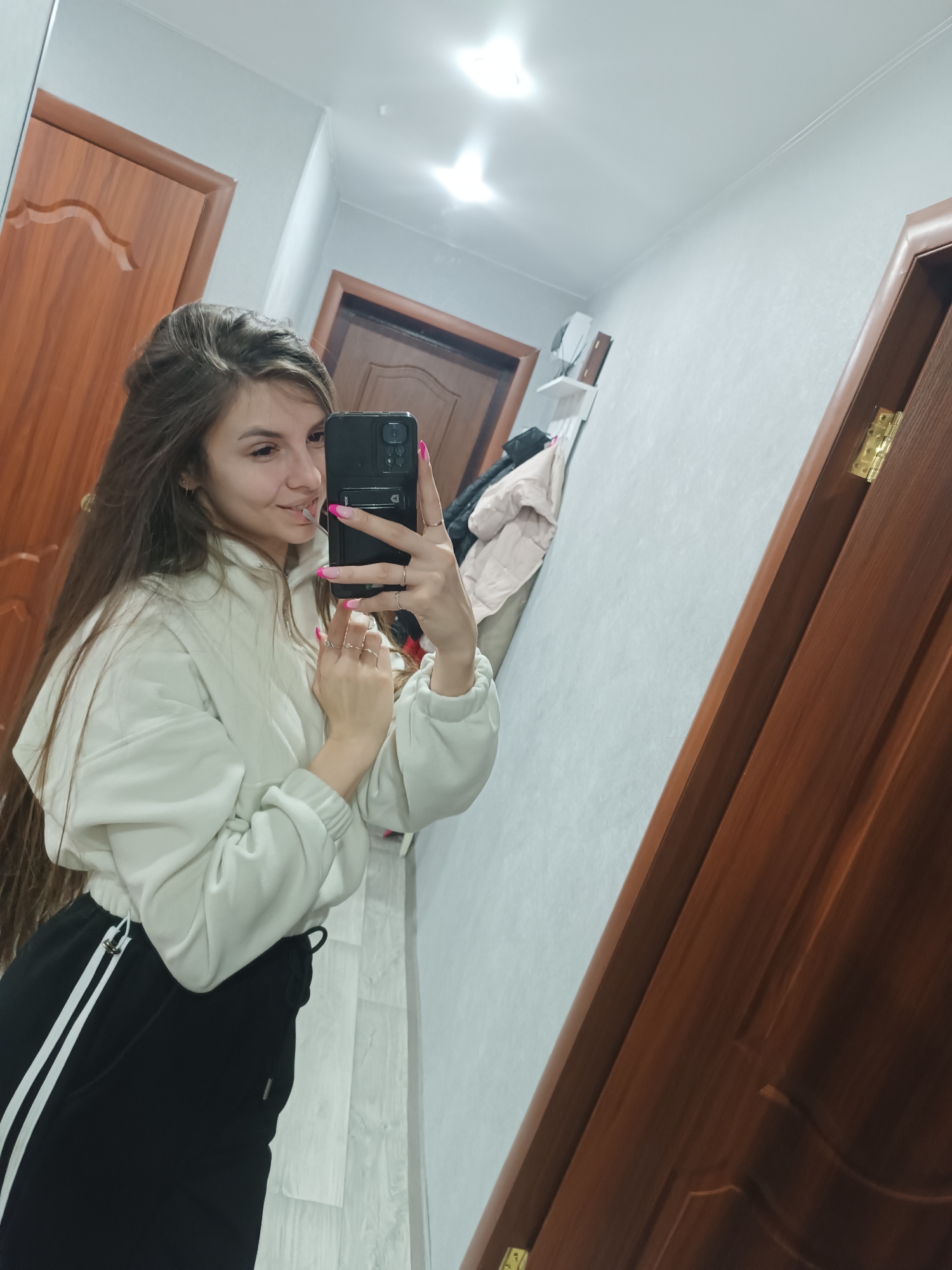 An indoor scene depicts a young adult female reflected in a mirror, taking a selfie with a dark-colored smartphone. The female has long, dark brown hair, is smiling, and wears a light-colored (off-white) cropped hooded sweatshirt and black athletic pants with white stripes on the left leg. Her fingernails are painted pink, and she wears several rings on her right hand. The environment reflected in the mirror appears to be a hallway or room within a private residence in Unknown, Unknown. Visible elements include two dark brown wooden panel doors, light grey walls, a white ceiling with a single bright light fixture, and a wall-mounted white shelf with items and a light-colored jacket hanging below it. The door on the right is slightly ajar, revealing a brass hinge. The flooring is light-colored, possibly laminate.