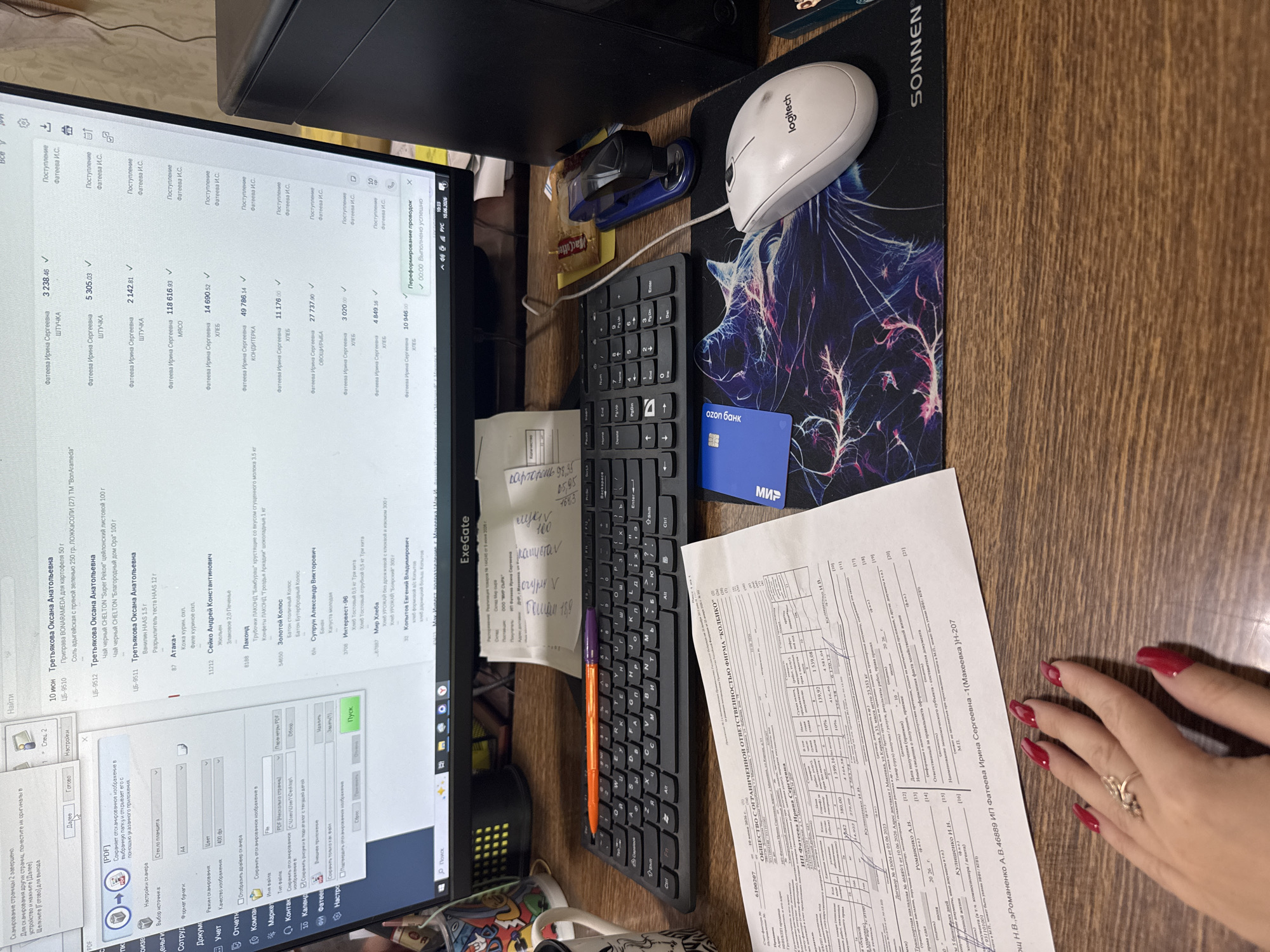 The image shows a desk with a computer monitor, keyboard, mouse, and documents. The computer monitor displays a software interface with lists of names and financial transactions. There are also several paper documents scattered on the desk, including what appears to be invoices or receipts. A stapler and a blue credit card are also visible. The scene suggests a workspace where financial or administrative tasks are being performed. The exact location is unknown, but it appears to be an indoor setting.