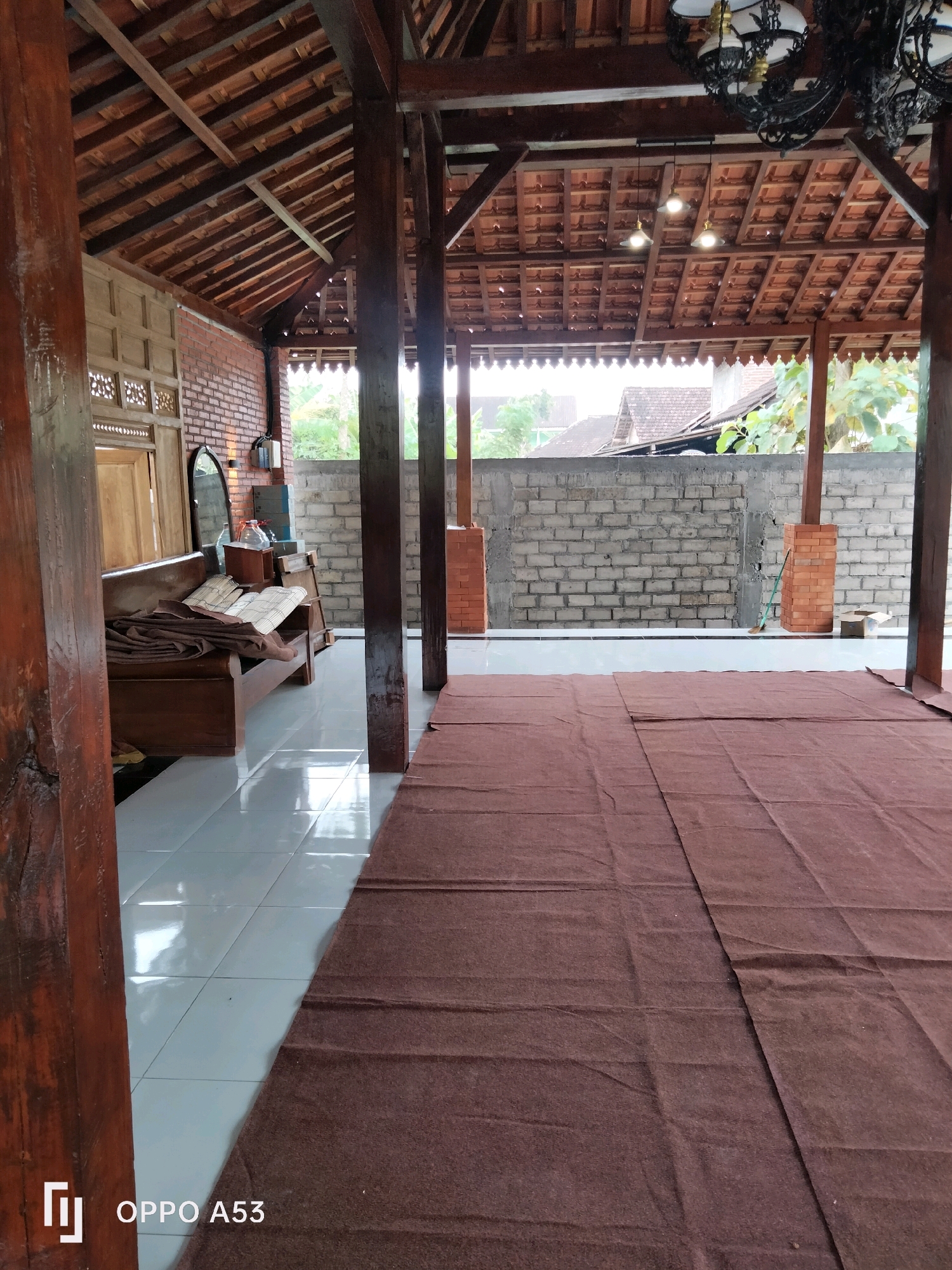 The image depicts an outdoor covered area with a traditional Indonesian architectural style, possibly a veranda or pavilion. The structure features wooden pillars and a tiled roof supported by wooden beams. The floor is tiled with large, light-colored square tiles. A large brown rug covers most of the floor in the foreground, with its edges visible on the right and bottom of the frame. In the background, a low brick wall separates the area from an outdoor space with some vegetation and buildings. To the left, a wooden bench with cushions and a mirror mounted on the brick wall are visible. The overall impression is of a functional, open-air space with a rustic, natural aesthetic. The image does not contain any people or identifiable landmarks that would allow for precise location within the city of Trenggalek, Indonesia.