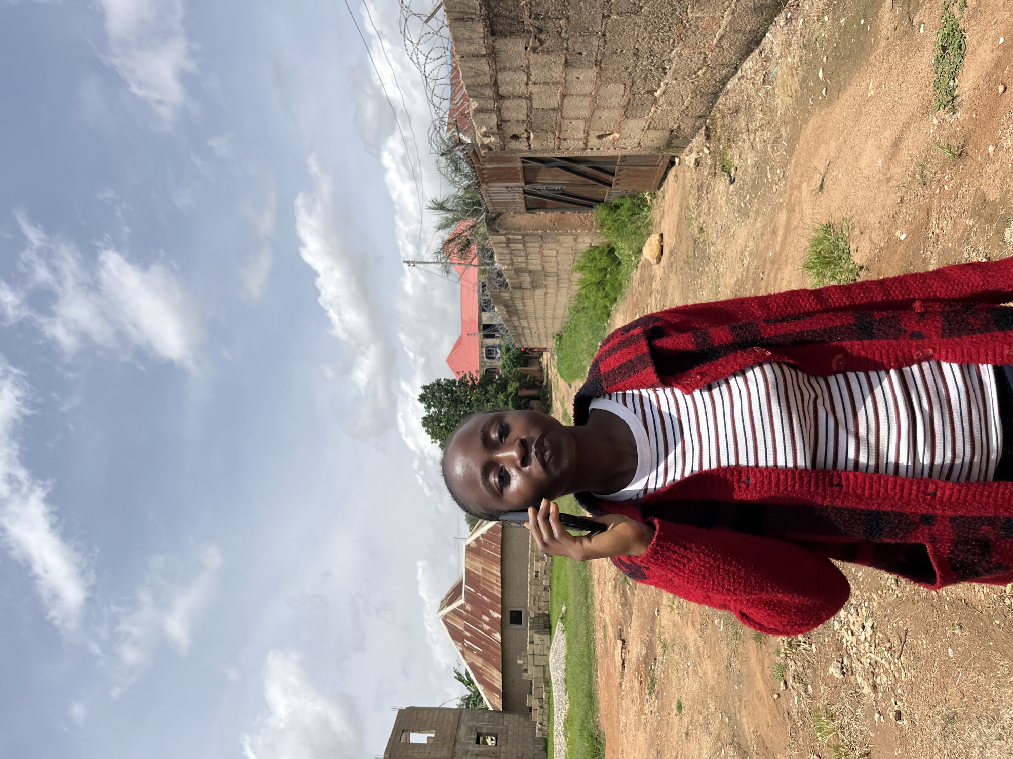 A young Black woman with short hair is standing outdoors in a dusty, unpaved area, holding a dark-colored mobile phone to her right ear. She is wearing a red cardigan with black plaid patterns over a white t-shirt with horizontal red and dark blue stripes. The background shows a rural or semi-developed environment in Bukuru, Nigeria. To the left, there is an unfinished brick building under construction with scattered bricks and a house with a corrugated metal roof. A tall, tan brick wall with barbed wire on top runs along the right side of the dirt path, and a rusted metal gate is visible within it. Further in the background, a building with a red roof is visible, surrounded by trees. The sky is partly cloudy with a light blue hue.