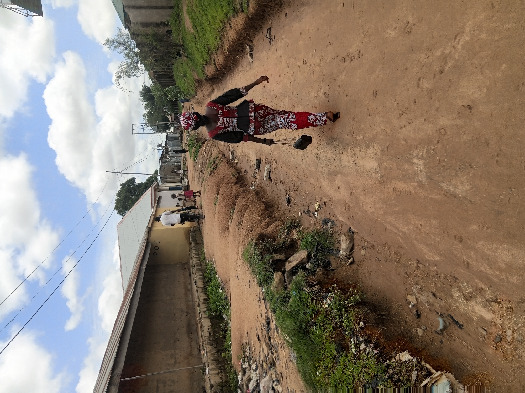 This image depicts a residential street scene in the city of Jalingo, Nigeria, viewed from slightly above and behind figures walking away from the camera. The setting is an unpaved, reddish-brown dirt road that appears worn and uneven, displaying tire tracks and numerous footprints.
In the foreground, a woman walks centrally on the dirt path, facing away from the observer. She is dressed in a matching ensemble consisting of a long-sleeved, peplum-style top, trousers, and a headwrap, all featuring a red, black, and white geometric pattern. She carries a small black handbag with a chain strap in her left hand and wears dark sandals.
To the immediate left of the road, a distinct eroded ditch filled with green vegetation and assorted litter, including plastic bags and debris, separates the pathway from a long, single-story building. This building features a corrugated metal roof and a tan-colored wall with the letters "POS" painted in black. Further down the street on this side, several children are visible, including one in a pink dress, near a man in a white t-shirt and dark trousers, all walking in the same direction.
On the right side of the road, a raised dirt embankment, partially covered with sparse green grass and scattered refuse, slopes upwards towards various simple buildings with corrugated roofs and concrete walls. A utility pole with power lines is visible on this side of the road in the mid-ground, alongside what appears to be an electrical transformer structure.
The background shows more residential structures, additional indistinct figures walking, and scattered trees under a bright, partly cloudy sky filled with white cumulus clouds. The overall environment suggests a semi-urban or rural residential area with basic infrastructure and visible evidence of open waste disposal along the roadside.