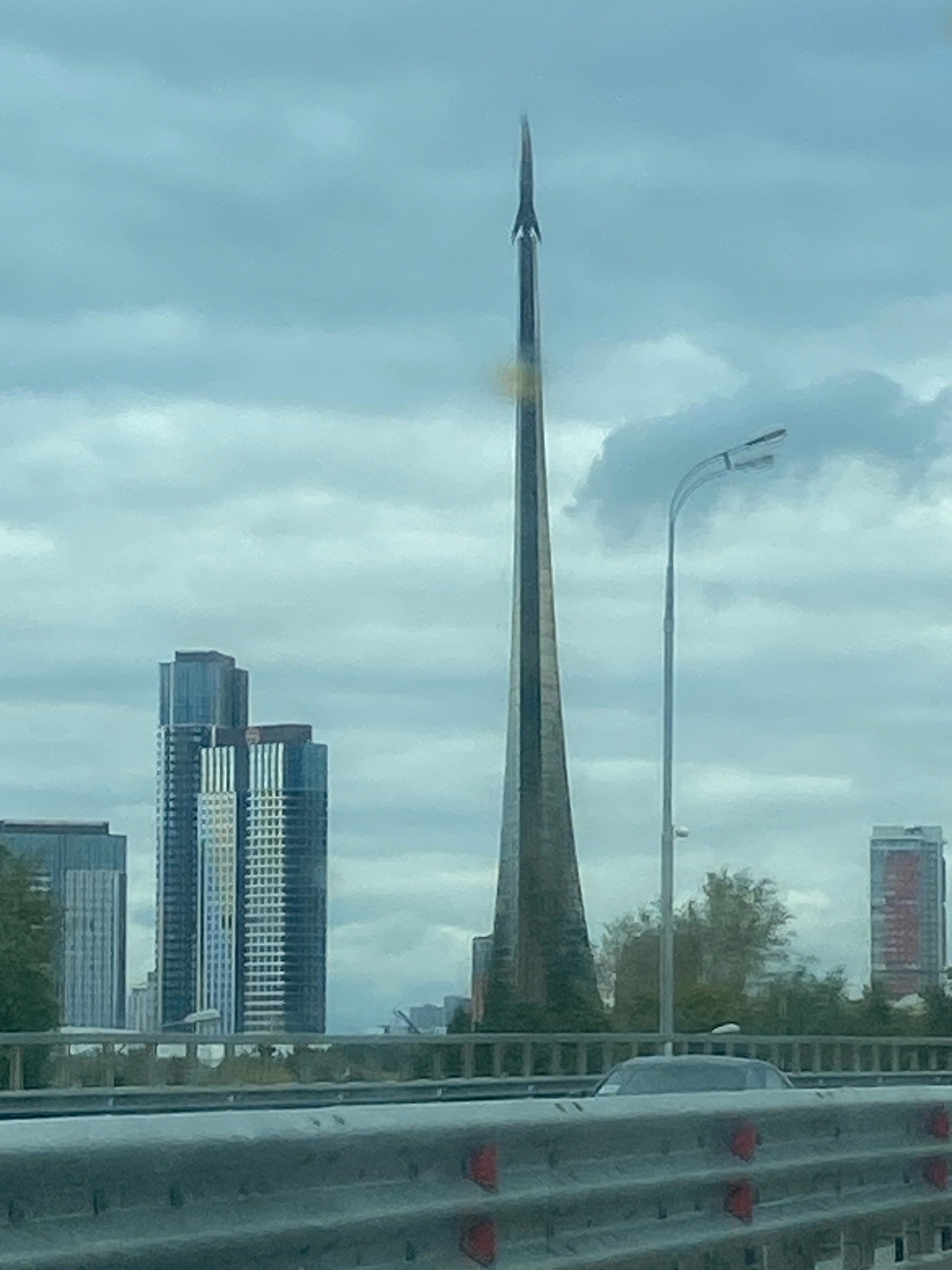 The image depicts the Ostankino Tower in Moscow, Russia, on a cloudy day. The tower, a prominent landmark, is seen in the center of the frame, its tall, slender structure tapering to a pointed spire. To the left of the tower, several modern skyscrapers are visible, characterized by glass facades and varied heights. A street light with two lamps stands to the right of the Ostankino Tower. In the foreground, a metal guardrail with red reflectors suggests a road or bridge setting. A dark-colored car is partially visible behind the guardrail. The sky is overcast with layers of clouds. The overall scene is urban, with a focus on the city's architecture and infrastructure under a somber sky.
