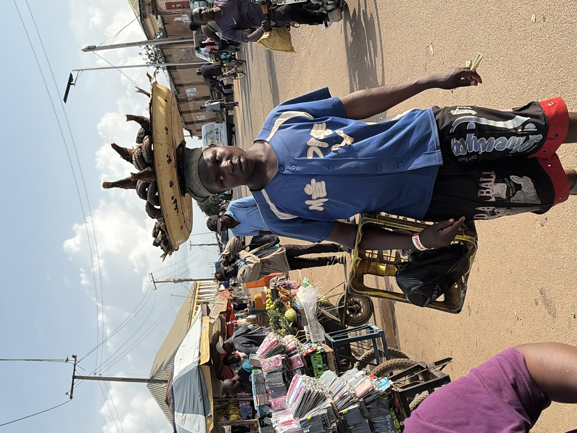 A man is walking down a dirt road in Gura Top, Nigeria, carrying a large basket of dried fish on his head. He is wearing a blue baseball jersey with white trim and black shorts. In his right hand, he holds a small bundle of sticks, possibly for sale. To his left, a cart is piled high with cell phone cases. In the background, other people are walking and riding bicycles. The sky is clear and blue, with a few white clouds.
