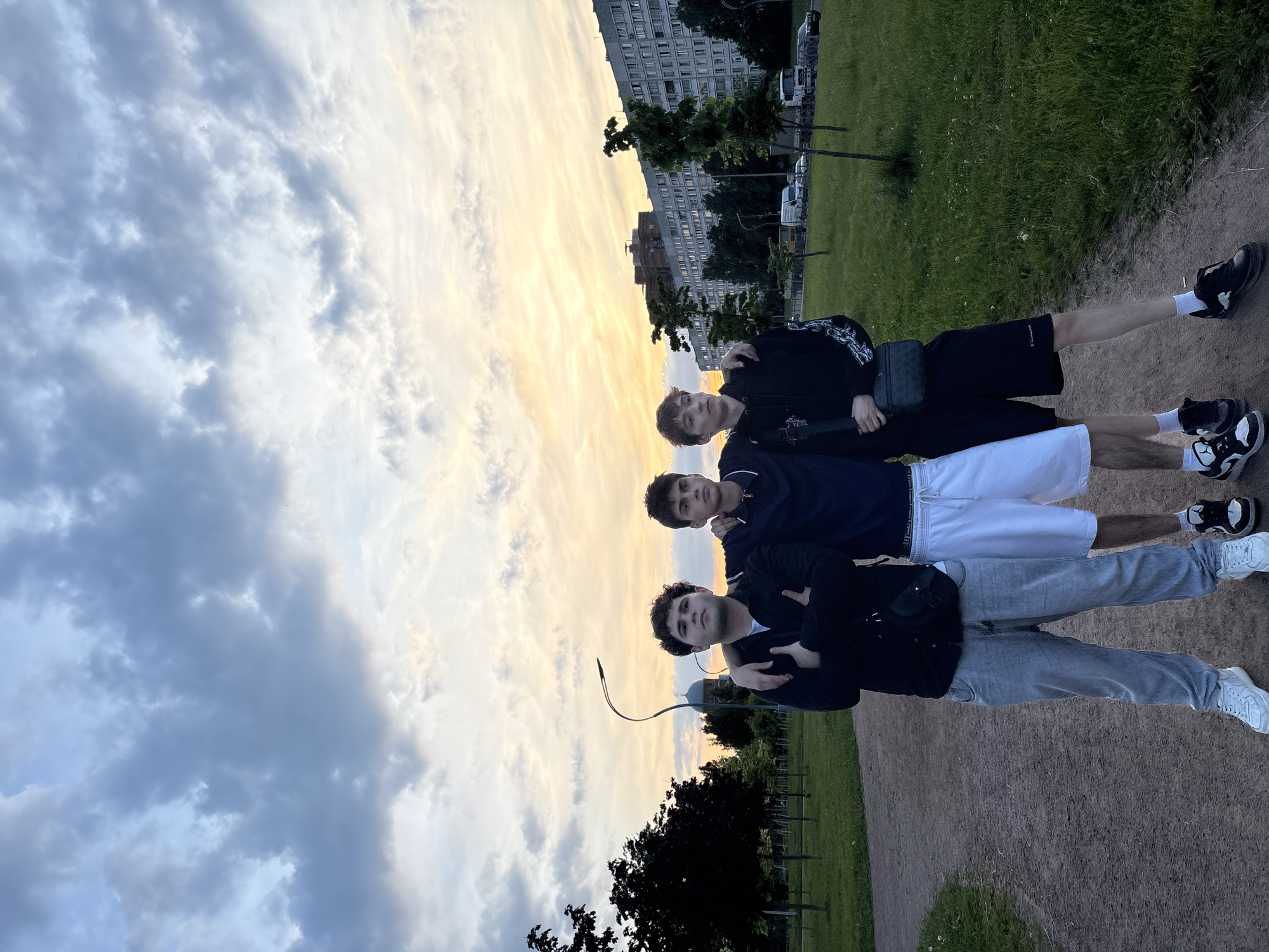 Three young men stand in a park on a dirt path with a grassy area to their right. The sky above is filled with dramatic clouds, with a sunset casting a warm glow. In the background, there are trees, a street lamp, buildings, and some vehicles. The man on the left is wearing a black jacket, grey pants, and white sneakers. The man in the middle is wearing a dark blue jacket, white shorts, and black and white sneakers. The man on the right is wearing a black hoodie, black shorts, and black sneakers. All three men have their arms around each other. The location is identified as Sinyavino, Russia.