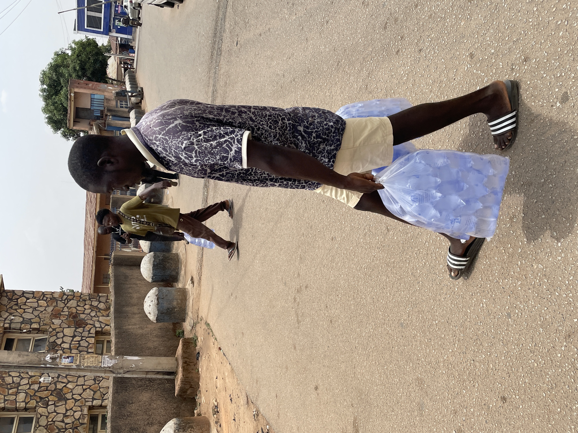 A medium shot depicts a street scene in Jos, Nigeria. A young man in a patterned, dark-colored shirt and light-colored shorts is walking down the street, carrying large, clear plastic bags filled with small, individually packaged water sachets. He is wearing striped sandals. Another individual is walking ahead of him, also carrying water sachets. Concrete barriers or bollards line the side of the road. Buildings, some constructed with stone, are visible in the background. The weather appears to be sunny.