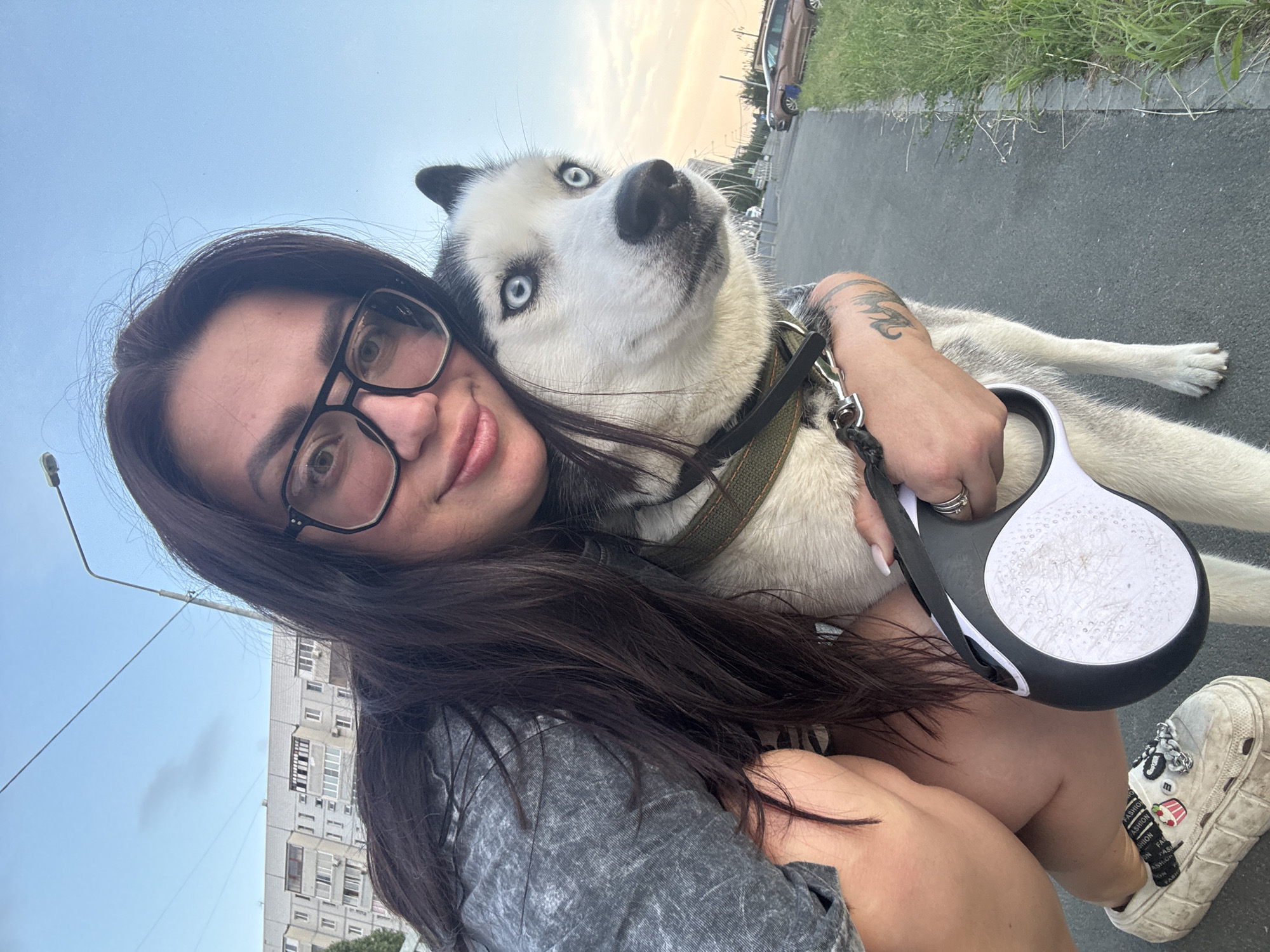 A woman with long dark hair and black-rimmed glasses takes a selfie with a husky dog on a sidewalk. She is wearing a gray t-shirt and denim shorts, and the dog is wearing a green collar. In the background, there are apartment buildings typical of Tolyatti, Russia, a street lamp, and parked cars. The sky is clear with a hint of sunset. The image captures a moment of companionship between the woman and her dog in an urban environment.
