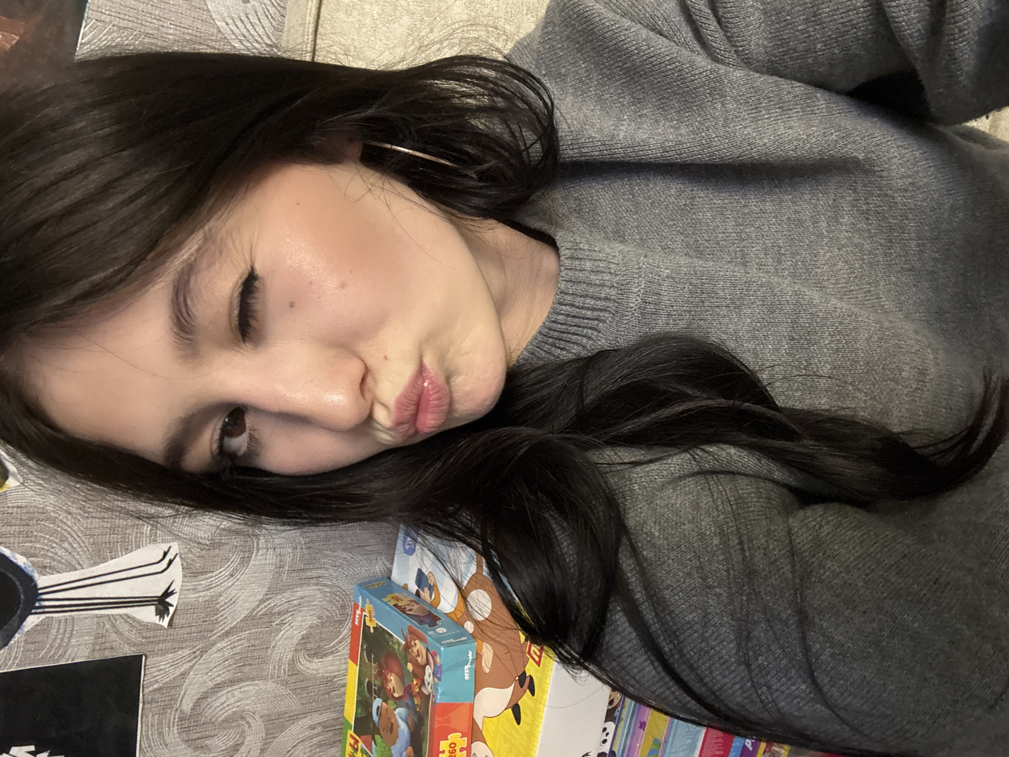 This image depicts an indoor scene, possibly in a residence located in the city of Фоки, Russia. The primary subject is a young adult female, positioned centrally and occupying the majority of the frame from the upper chest upwards. She has long, dark hair, styled down, and is wearing a grey, ribbed knit sweater. A thin, gold-colored hoop earring is visible in her right ear. Her right eye is closed in a wink, and her lips are pursed in a kissing gesture, directed towards the camera.
The background consists of a wall covered with light-colored wallpaper featuring a subtle, wavy or swirling pattern. Two cut-out figures are affixed to the wall on the upper left: one partially visible black shape and one white shape with black linear details resembling legs. To the lower left, a stack of colorful rectangular boxes, identified as "STEP Puzzle" brand, is visible. The topmost box features cartoon characters. A light-colored, upholstered furniture piece, possibly a couch or chair, is partially visible on the far right edge of the frame. Lighting appears uniform, suggesting an indoor artificial light source.