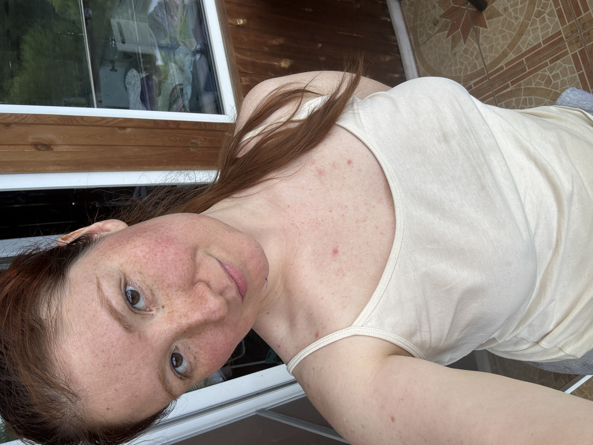A fair-skinned woman with red hair and freckles is seen from a high-angle, close-up perspective. She is wearing a light-colored tank top. The background includes a window, wooden paneling, and patterned floor tiles. The location can not be determined from this image.