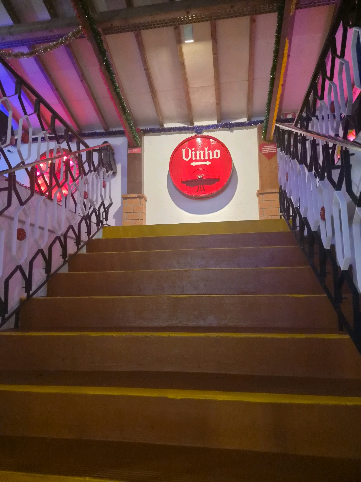 The image captures an interior view of a stairwell, likely in a commercial establishment, possibly a restaurant or bar, given the presence of a decorative sign. The stairs themselves are wide and appear to be made of wood, painted a bright yellow. The risers are a deeper brown color, and the edges of each step are also highlighted in yellow.
To the left and right of the staircase, there are decorative railings. The left railing features an ornate black metal design with white accents and some red decorative elements visible. Further up the left side, a bright red, circular sign with the word "Vinho" (Portuguese for "wine") and an arrow pointing left is prominently displayed on a white wall. Above the sign and railings, the ceiling consists of exposed wooden beams with some decorative Christmas tinsel strung across them. Red and purple lighting illuminates the ceiling area, casting a vibrant glow.
The right side of the staircase also has a similar ornate black metal railing with white accents. Above the railing, a portion of the wall is visible, with a small brick structure and another section of the white wall.
The overall impression is one of a festive and possibly rustic ambiance. The image is focused on the staircase, leading upwards into a brightly lit and decorated space. There are no people visible in the image. The scene suggests a location that embraces a lively or celebratory atmosphere.
It is not possible to determine the specific location within Darque, Portugal, from the image alone. However, the presence of Portuguese text on the sign strongly indicates the location is in Portugal.