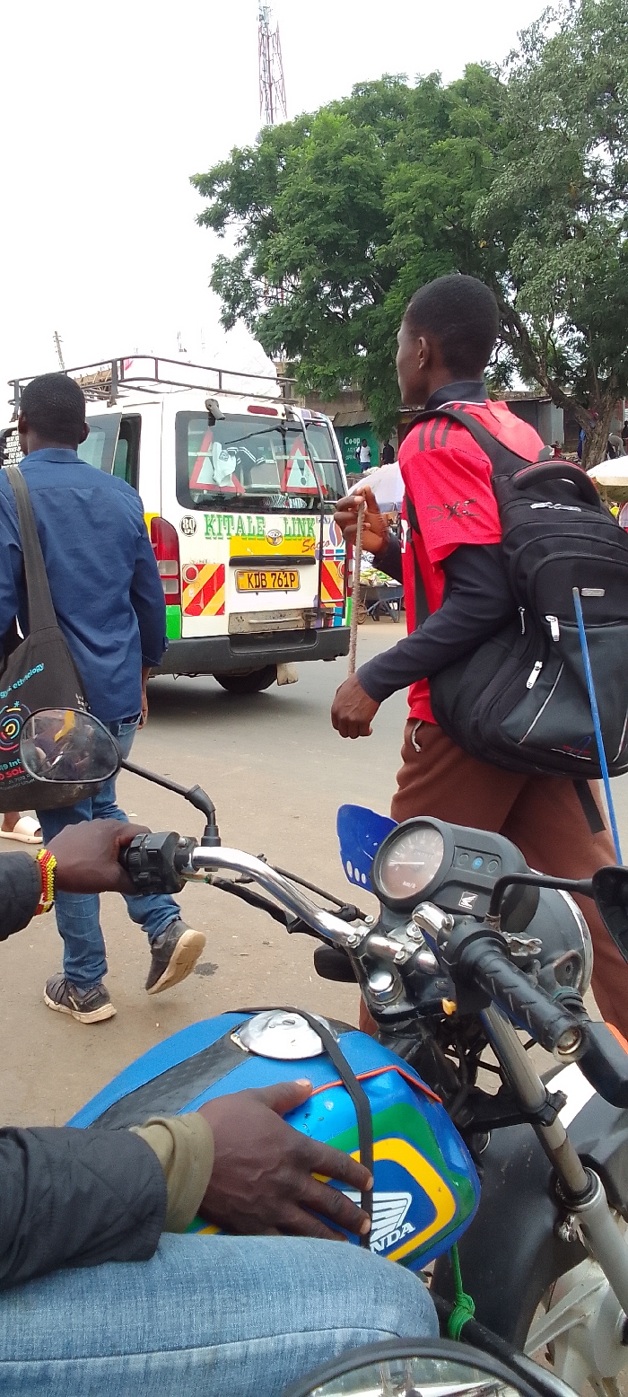 This daytime image captures a street scene in Kakunga, Kenya, under what appears to be an overcast sky, indicated by the diffused lighting. In the immediate foreground, a person is seated on a blue Honda motorcycle, with their hands on the handlebars and resting on the fuel tank, wearing blue jeans. A beaded bracelet adorns their left wrist.
In the midground, a white passenger van, typical of a Kenyan matatu, is prominently featured. Its rear displays "KITAL LINK SACCO" and the Kenyan license plate "KDB 761P", along with several orange triangular warning stickers on its rear window. To the left of the van, a man in a blue shirt walks away from the viewer, carrying a black shoulder bag with a colorful design that includes a partially visible word, "technology". To the right of the van, another man, wearing a red and black shirt and brown trousers, walks with a black backpack. He holds a long, thin, rope-like object in his right hand.
The background includes dense green trees and a tall communication tower reaching into the sky. The scene depicts typical daily activities on a public street, with people moving and vehicles present. The visible text includes "KITAL LINK SACCO" and "KDB 761P" on the matatu, and "HONDA" on the motorcycle's tank. The setting reflects a bustling area in a Kenyan town.